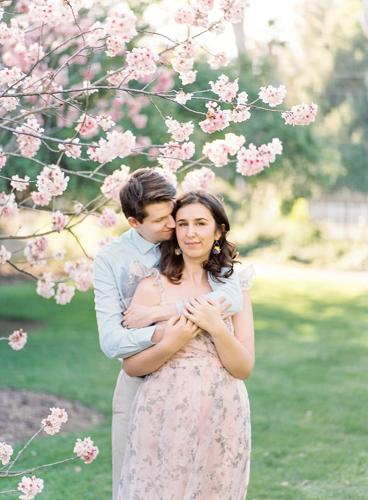 Cherry Blossom Photoshoot ideas featuring a couple embracing under blooming cherry blossom branches, with the man hugging from behind as soft sunlight filters through the petals for a romantic, intimate moment