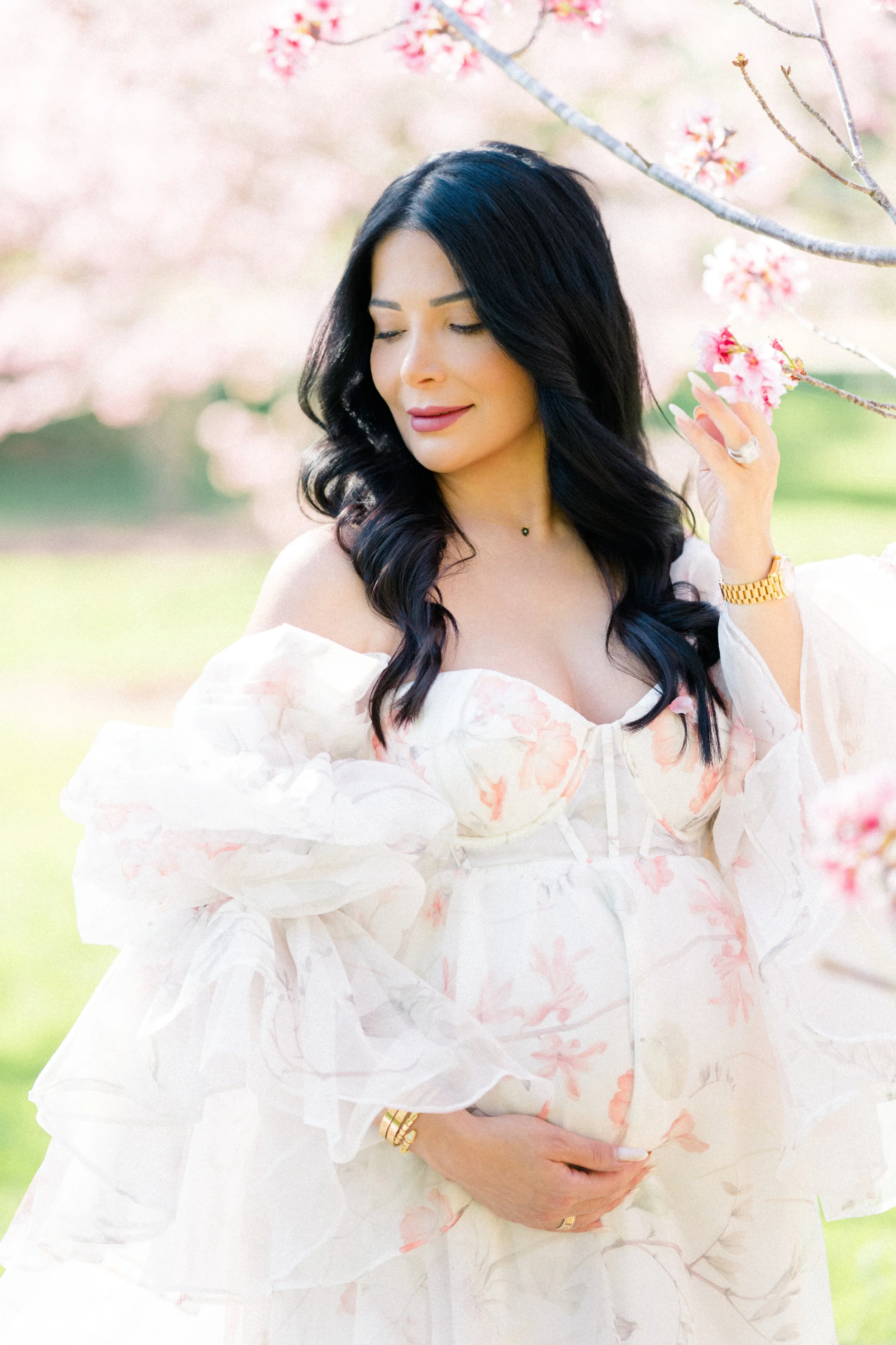 Cherry blossom Maternity photos of an expecting mother in a soft, flowing floral gown gently holding her baby bump while touching delicate pink blossoms in warm natural light.