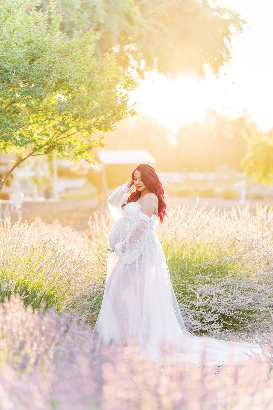 Pregnant woman in a flowing white gown standing in a lavender field at golden hour during a dreamy outdoor maternity photoshoot, surrounded by soft purple blooms and warm sunset light.