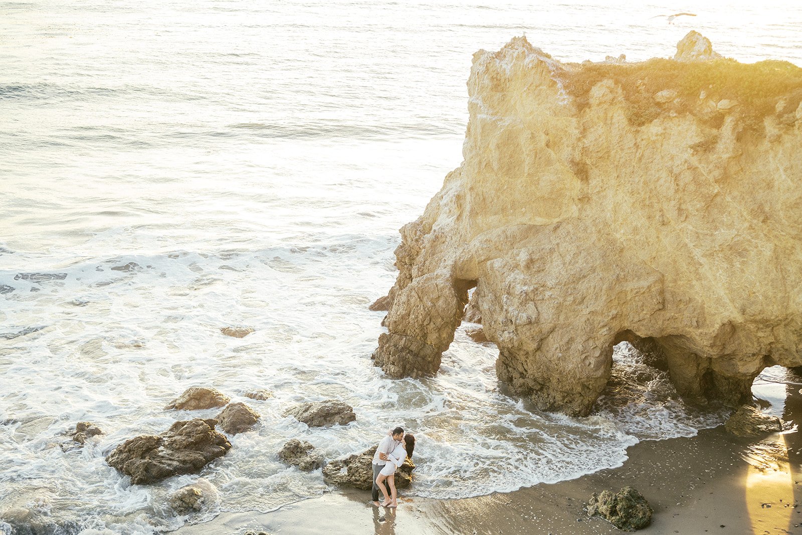A couple shares a romantic moment at the base of a golden sea cliff as gentle waves wash around them, the late-afternoon sun illuminating the rocks at a Malibu beach. Holiday Date Night Ideas.