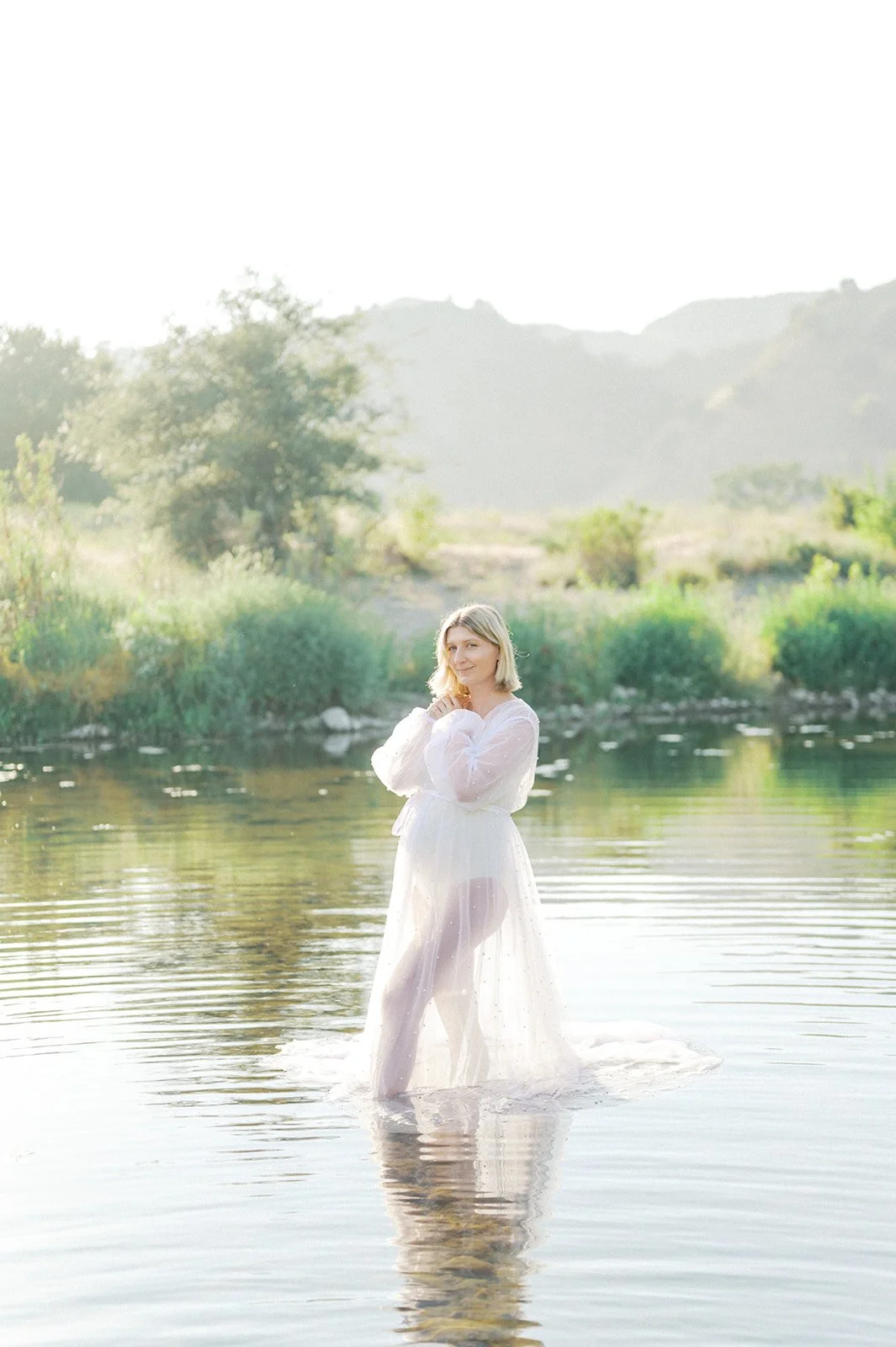 Pregnant woman stands gracefully in a shallow creek wearing a sheer, flowing gown, surrounded by soft morning light and lush green hills in the background.