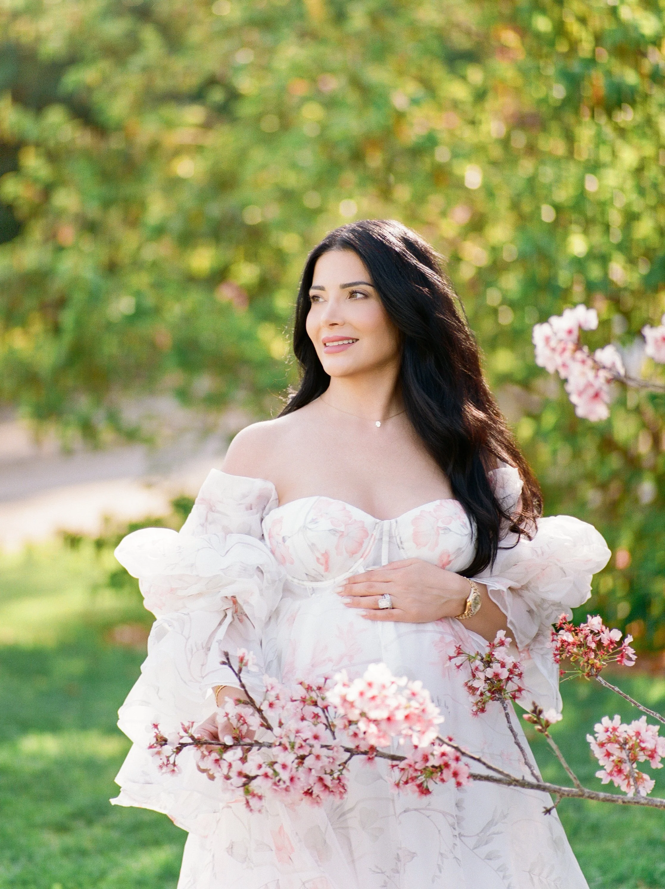 Cherry blossom Maternity photos of an expecting mother in a soft floral gown gently holding her baby bump while standing among blooming pink branches in warm, natural spring light.