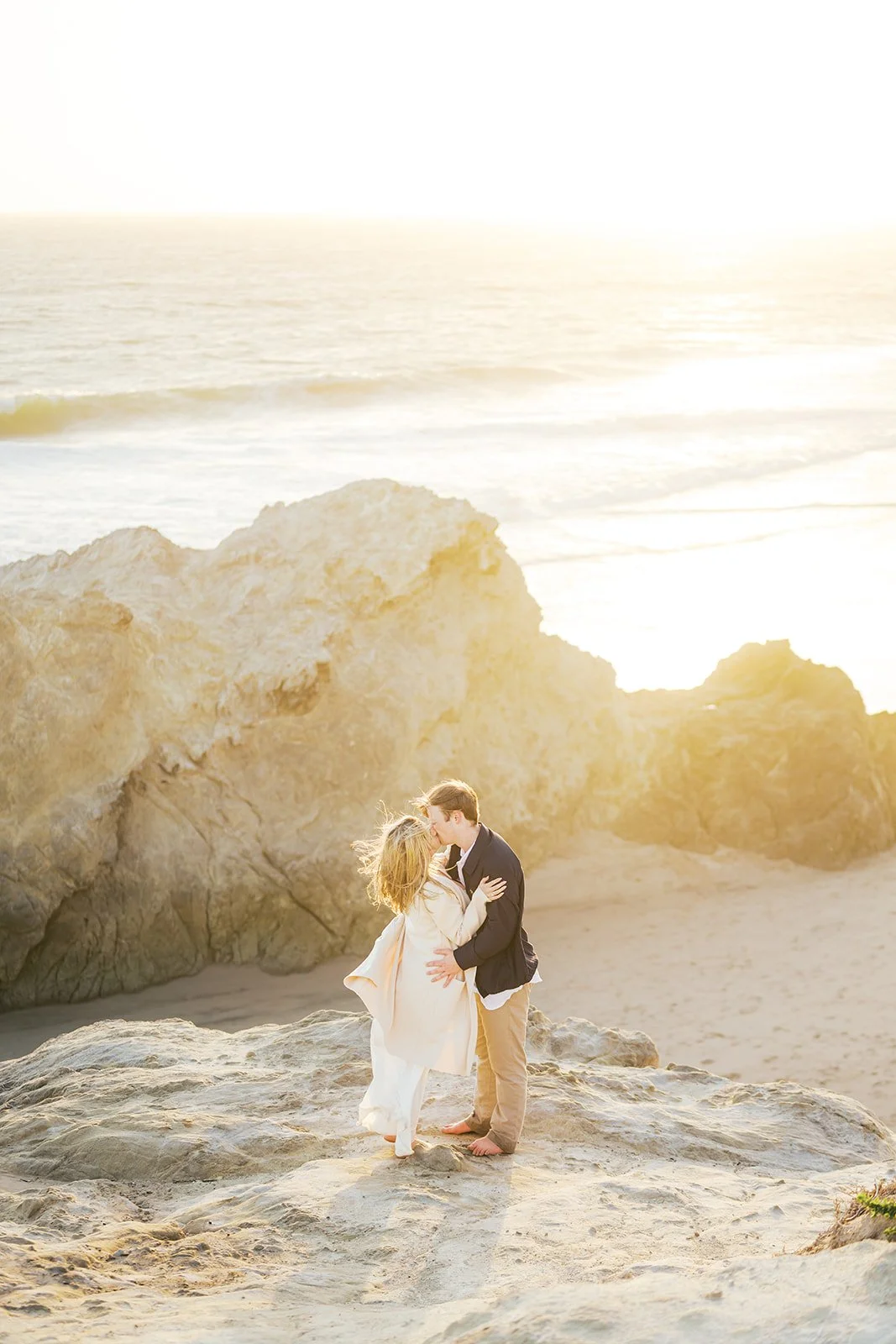 Summer Engagement Photos in Malibu featuring an engaged couple kissing on rocky cliffs overlooking the beach and Pacific Ocean during golden hour.