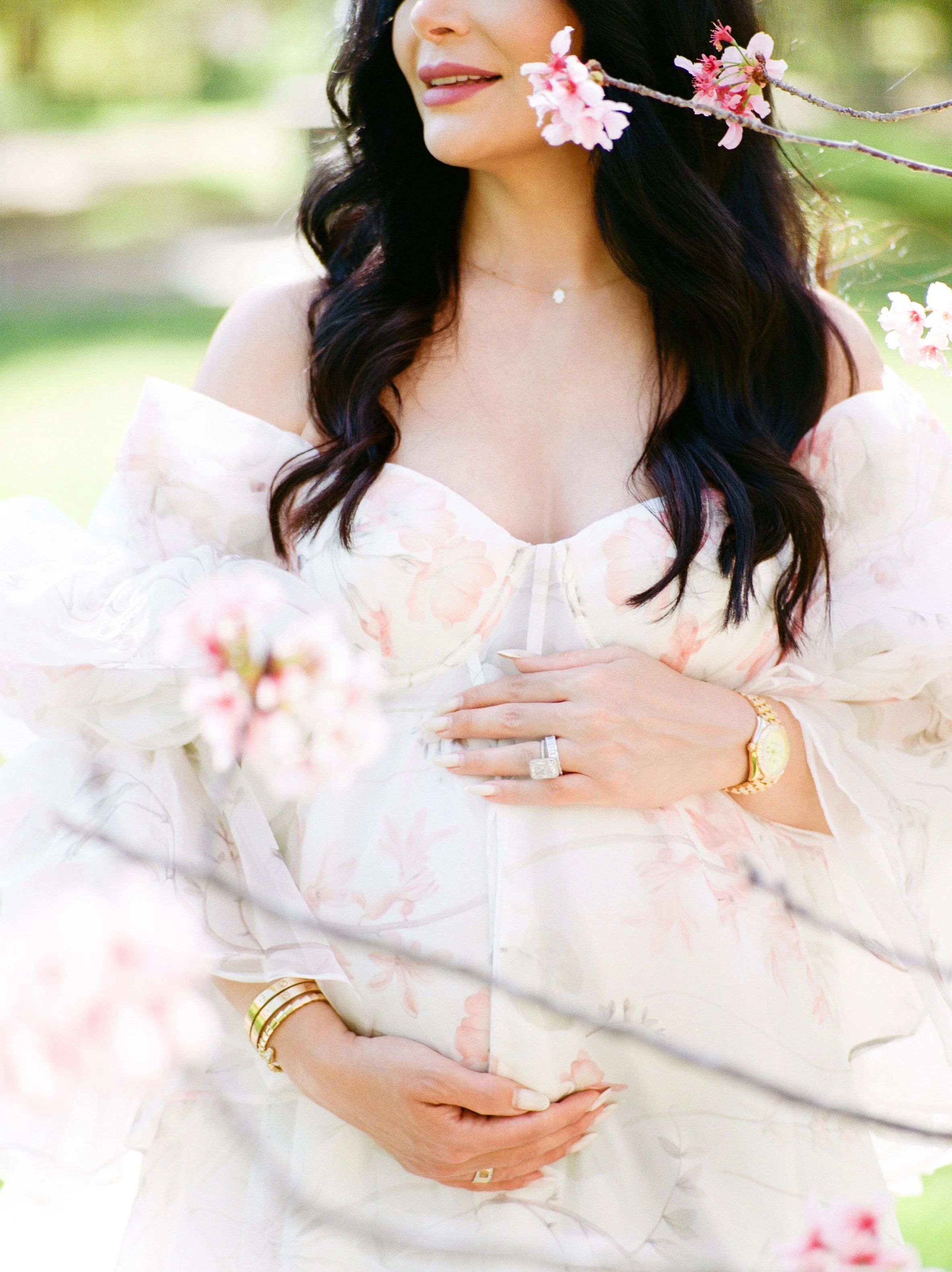 Cherry blossom Maternity photos close-up of an expecting mother in a soft floral gown gently cradling her baby bump, framed by delicate pink blossoms in soft natural light.