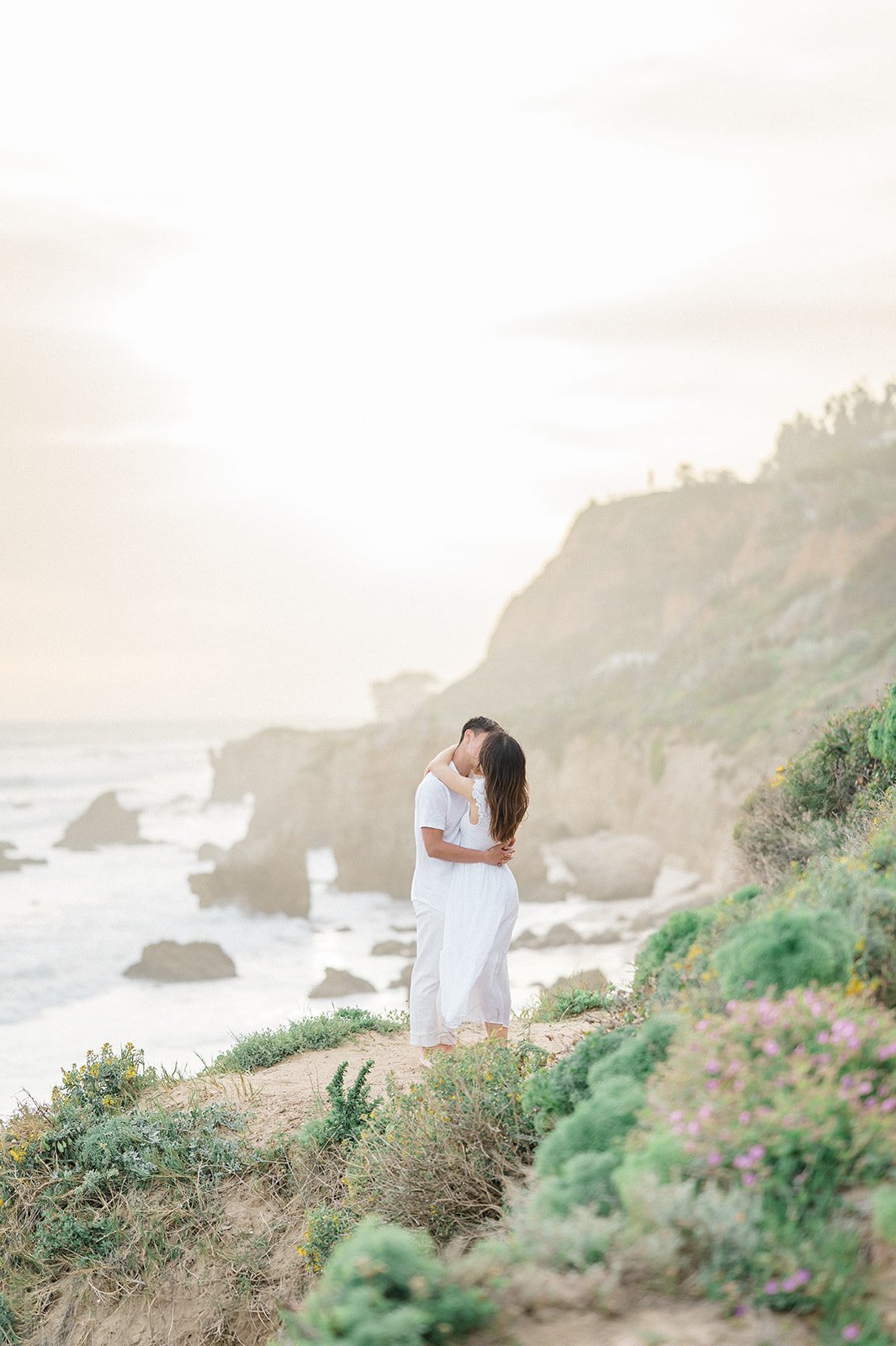 Summer Engagement Photos in Malibu featuring a couple kissing on a coastal bluff overlooking El Matador Beach, surrounded by wildflowers and dramatic ocean cliffs.
