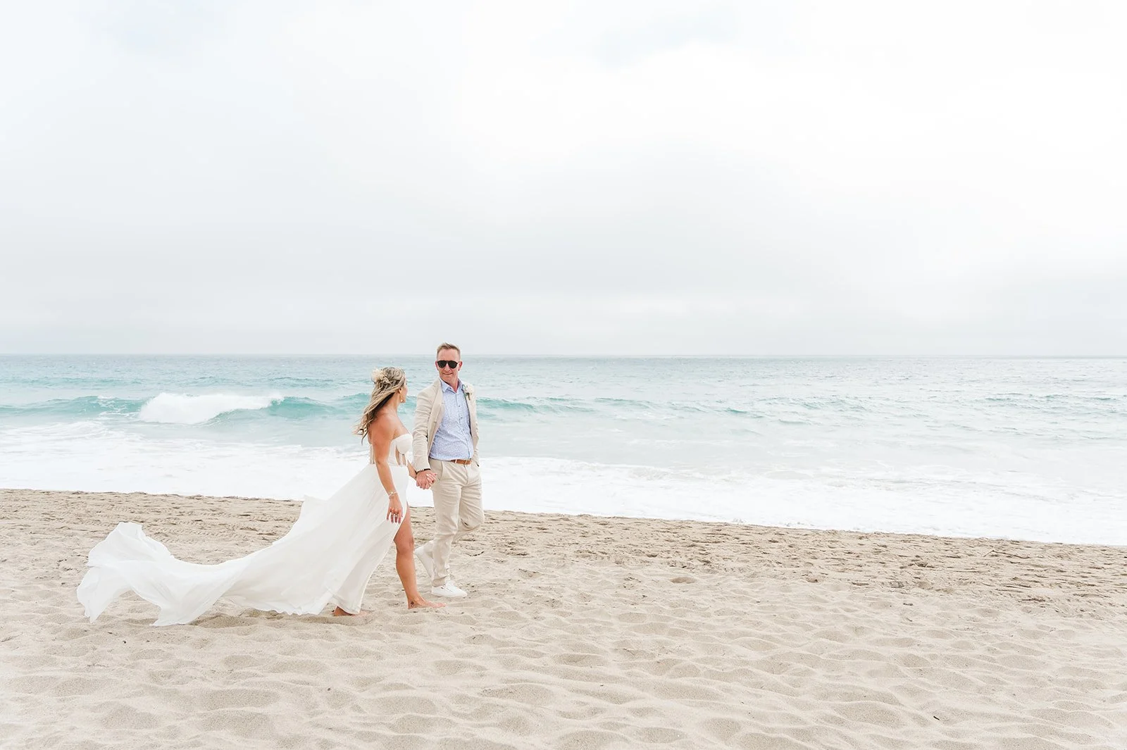 Bride and groom walk barefoot along a sandy beach, holding hands and smiling against a misty ocean backdrop. This relaxed coastal setting captures the carefree romance of one of the Best Places to Elope 2026.