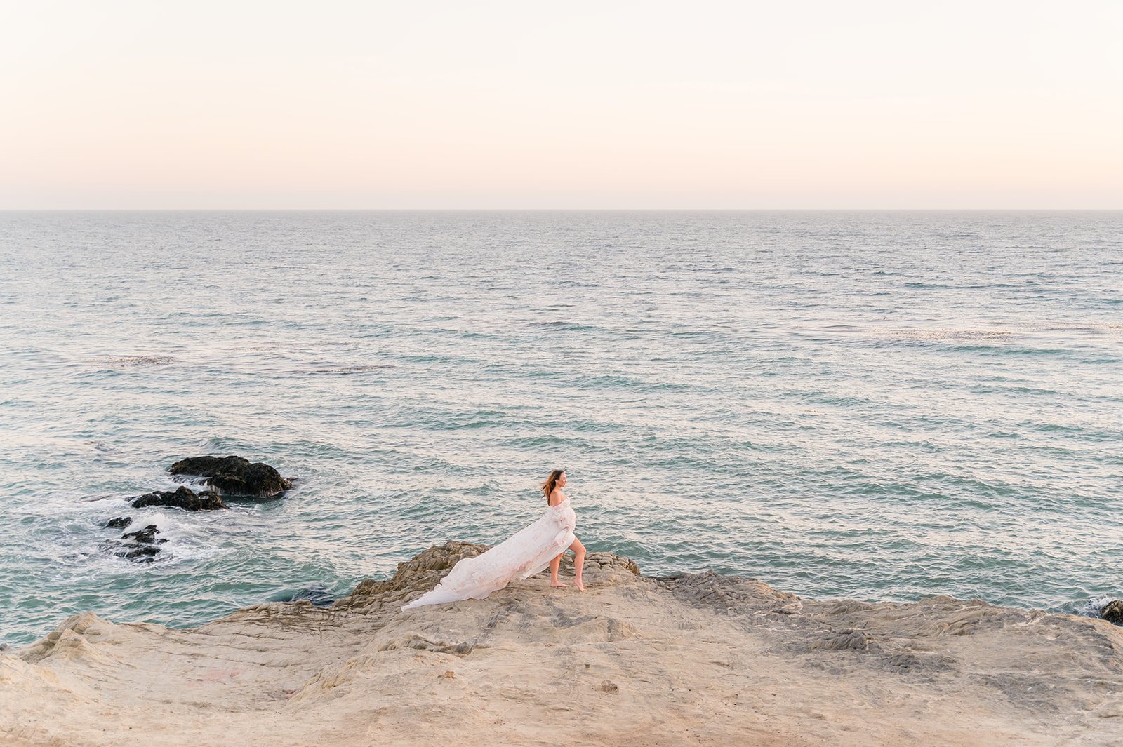 Summer Maternity Photos of an expecting mother in a flowing pink gown standing on coastal rocks overlooking the ocean during a pastel sunset.