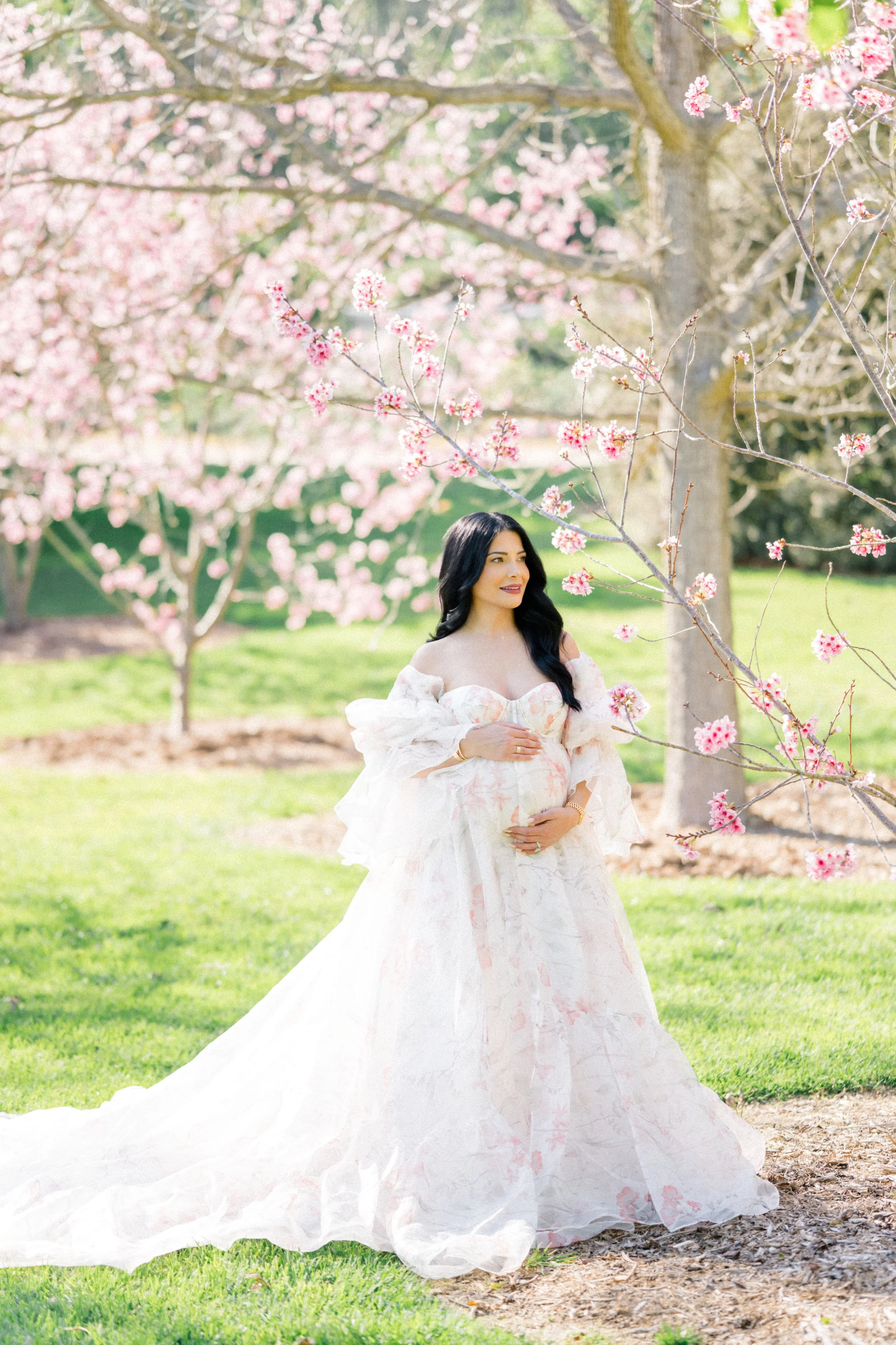 Cherry blossom Maternity photos of an expecting mother standing beneath blooming pink trees, wearing a flowing floral gown and gently holding her baby bump in soft, airy spring light.