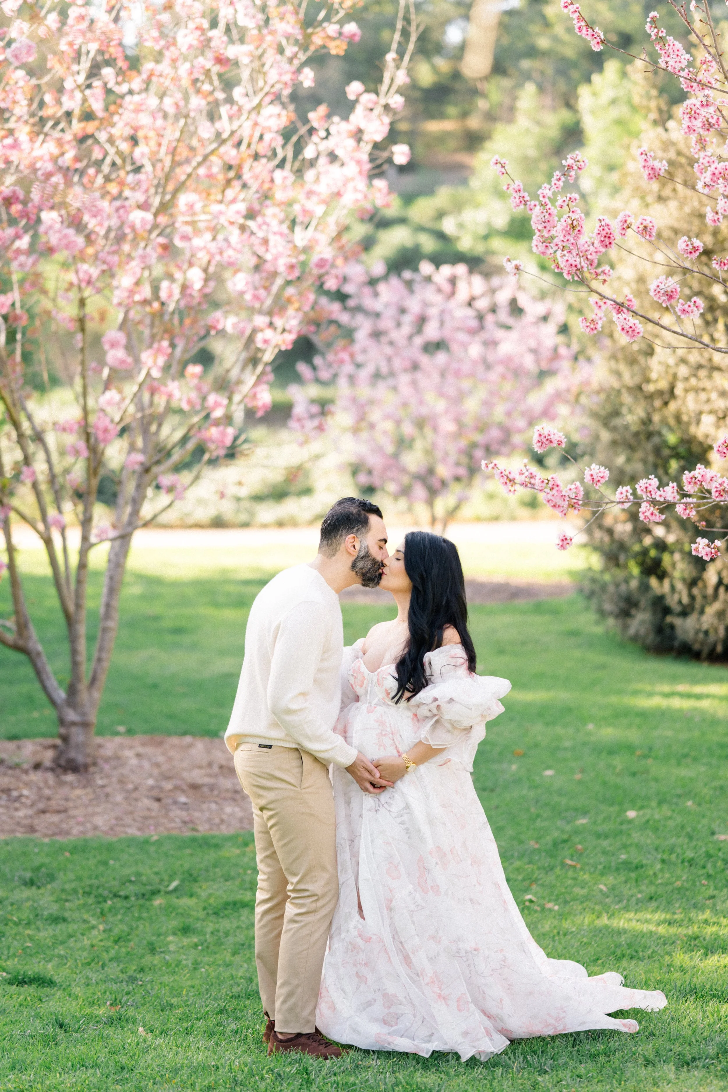 Cherry blossom Maternity photos of an expecting couple standing beneath blooming pink trees, sharing a kiss while gently holding the baby bump in soft, natural spring light.