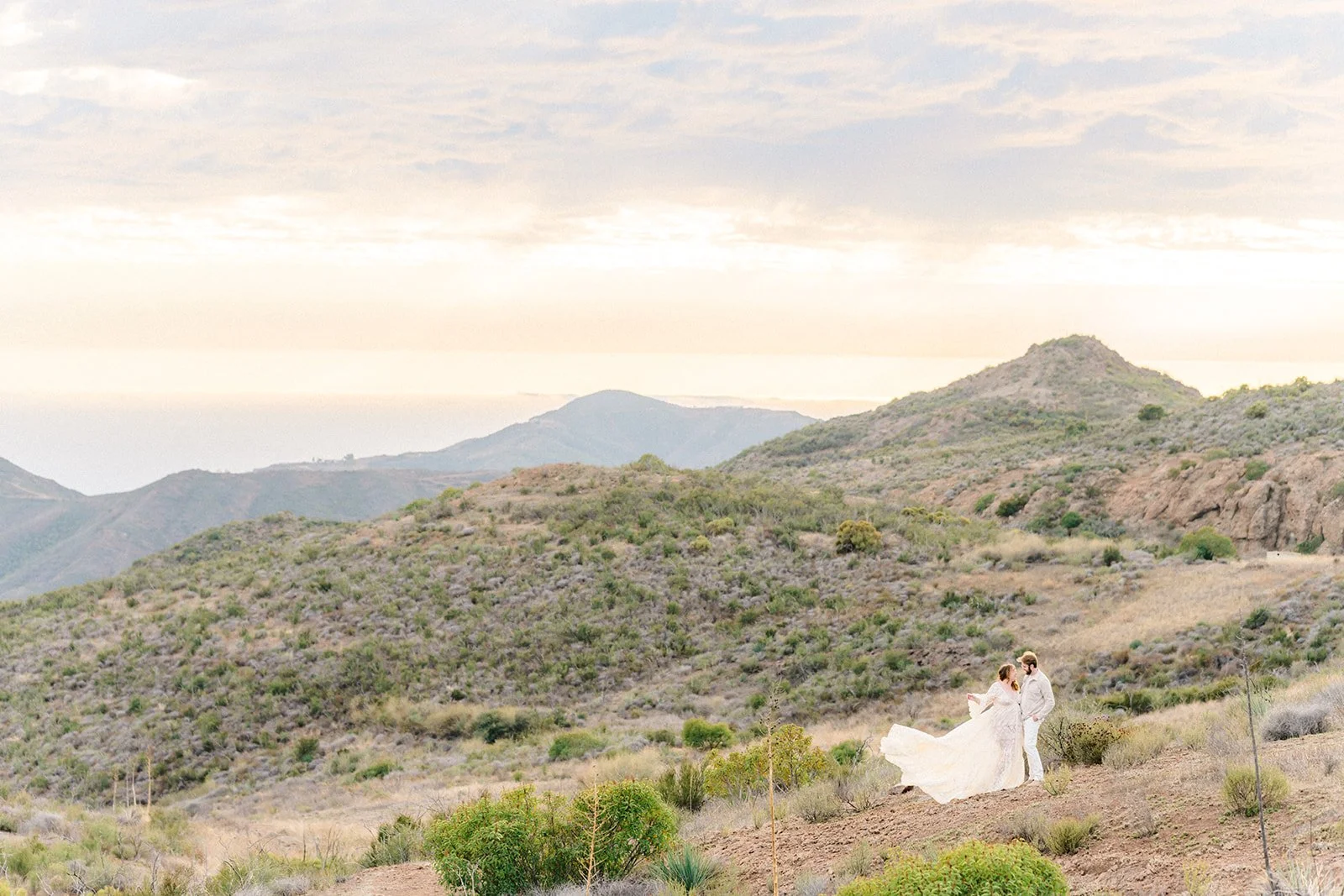 Summer Engagement Photos in Malibu featuring a couple embracing in the Santa Monica Mountains with rolling hills and the Pacific Ocean in the distance.