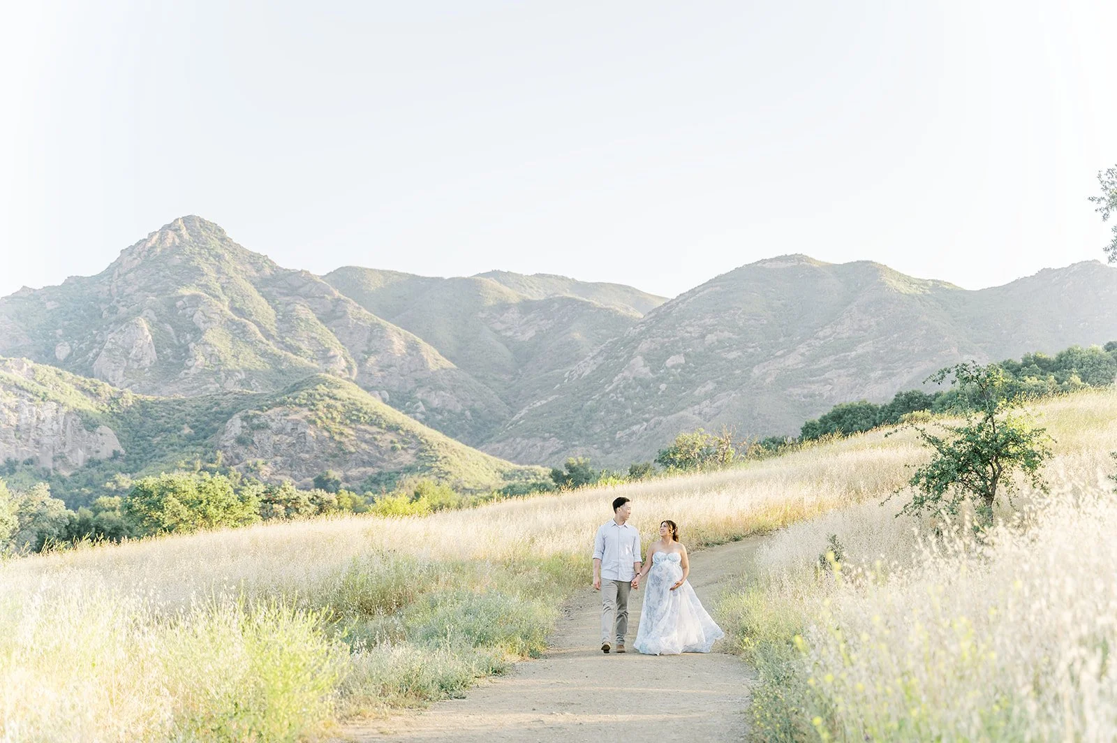 Summer Maternity Photos of an expecting couple walking through golden fields with mountain views in the background during a warm California sunset.