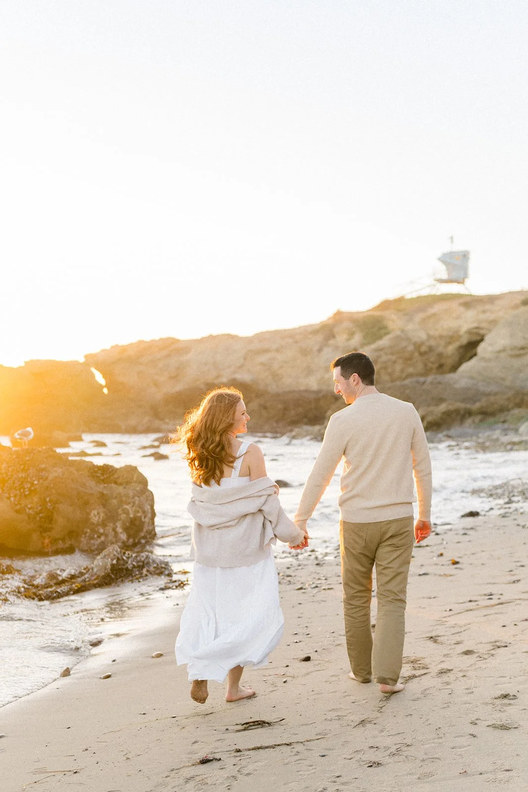 Summer Engagement Photos in Malibu featuring a couple holding hands and walking barefoot along a rocky beach at golden hour.