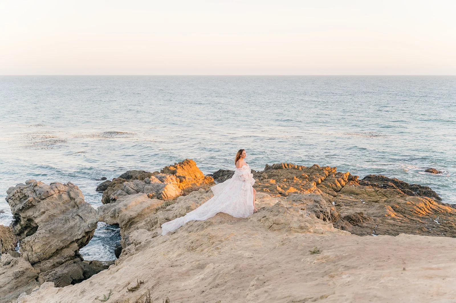 Summer Maternity Photos of an expecting mother in a flowing floral gown standing on rocky coastal cliffs overlooking the ocean during golden hour.