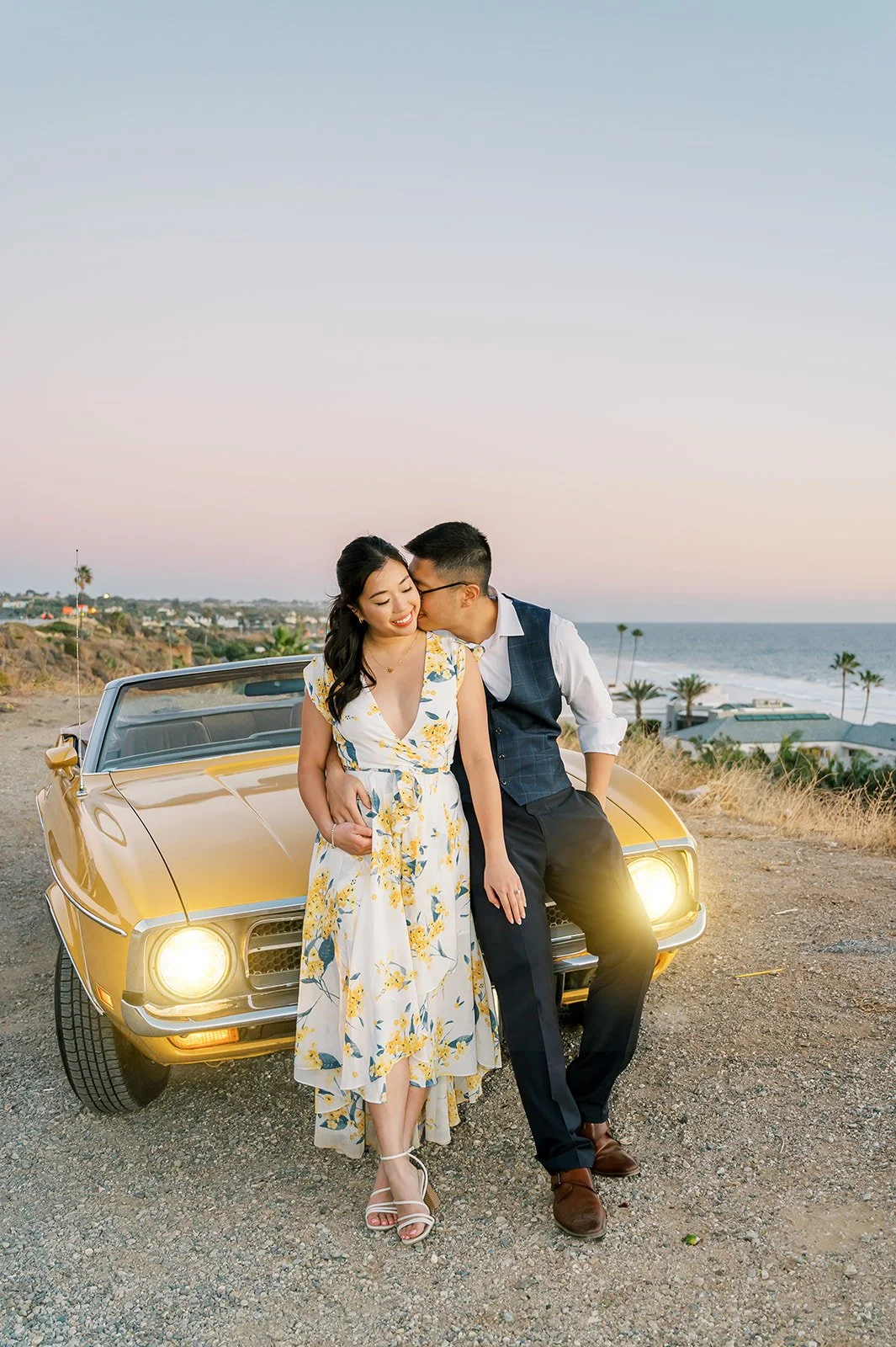 Summer Engagement Photos in Malibu featuring a couple cuddling beside a vintage yellow Ford Mustang convertible with the Pacific Ocean and pastel sunset sky in the background.