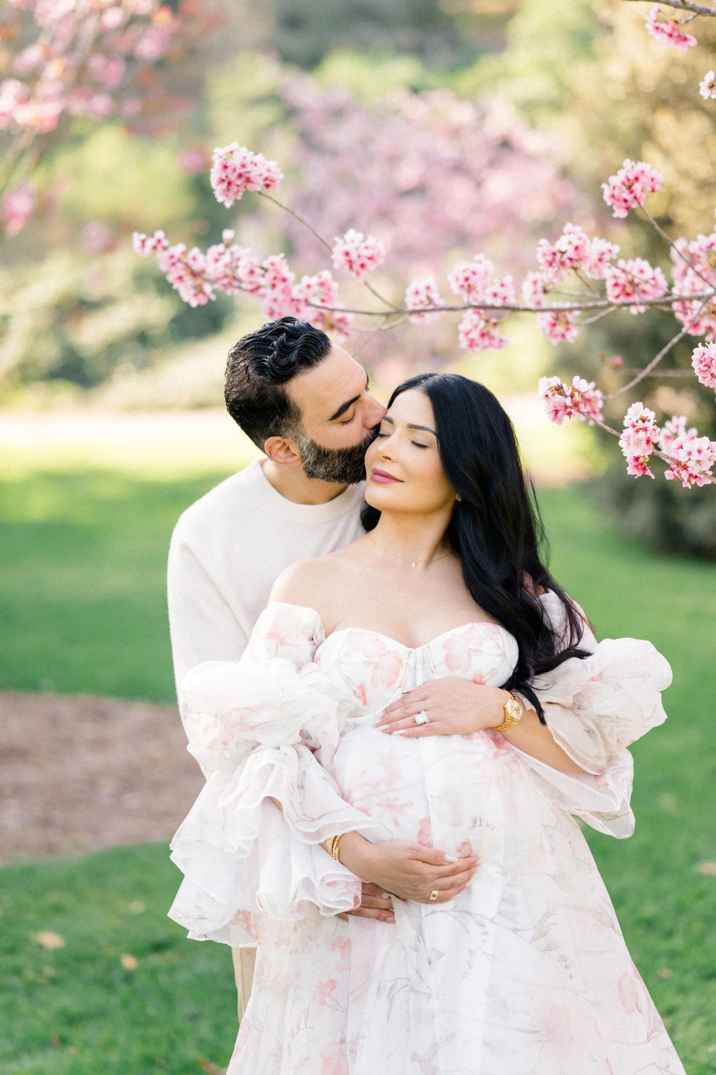 Cherry Blossom Photoshoot ideas featuring a maternity session with a couple embracing beneath blooming cherry blossom branches, as the partner gently kisses her cheek while she cradles her belly in soft spring light