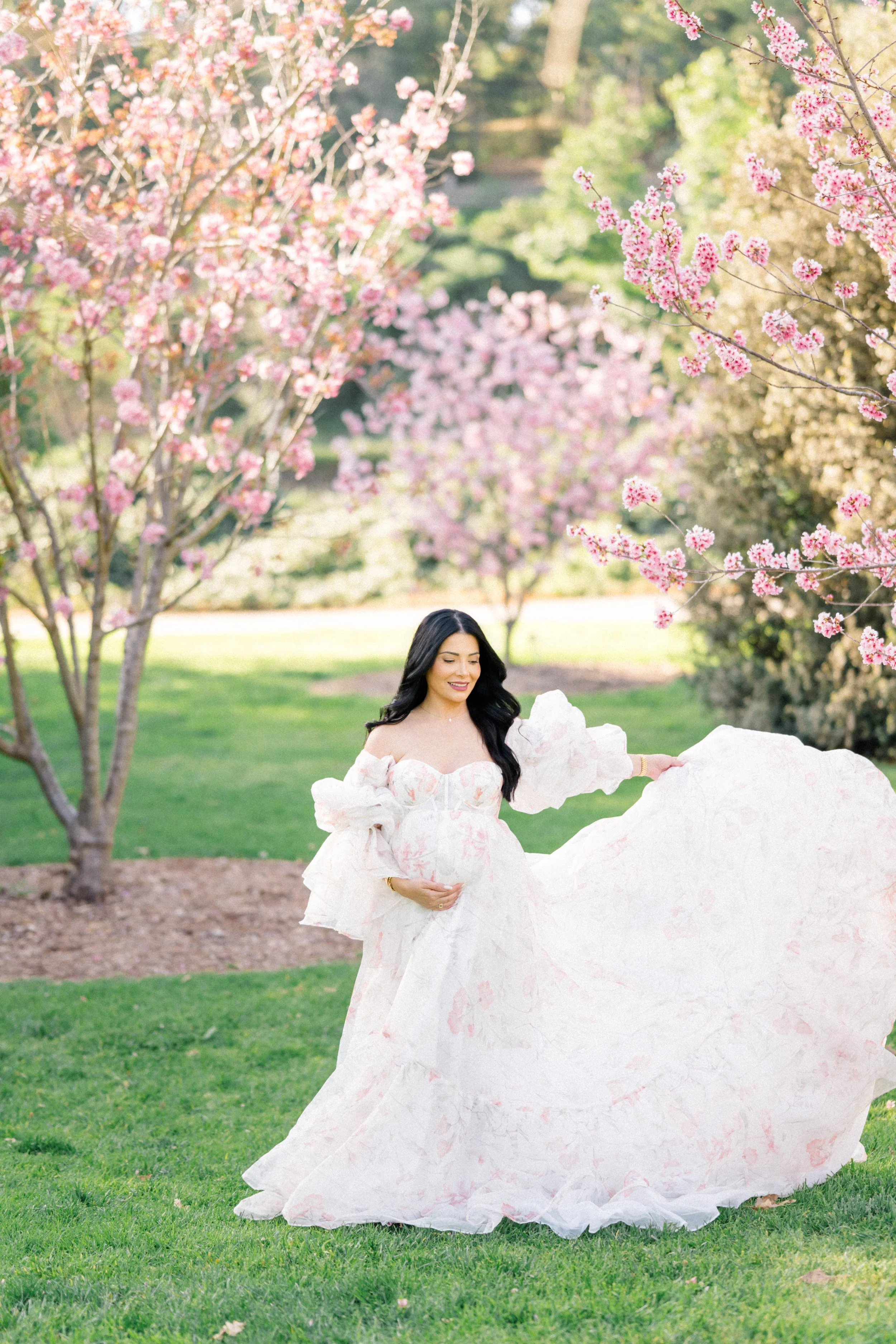 Cherry Blossom Photoshoot ideas featuring a maternity session with a woman in a flowing dress gently holding her belly while lifting the fabric, surrounded by blooming cherry blossom trees in soft spring light