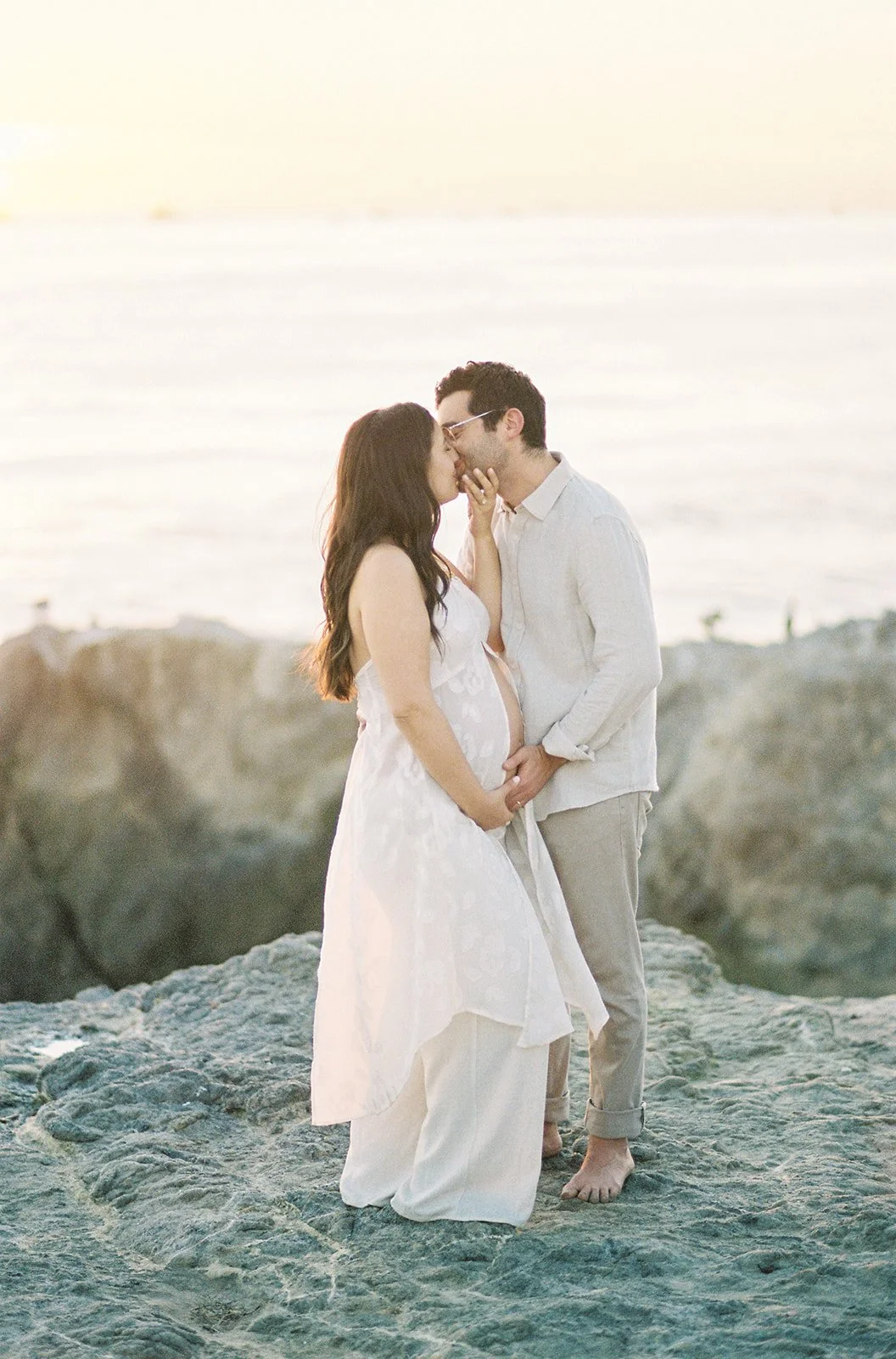 Summer Maternity Photos of an expecting couple sharing a kiss on coastal rocks at sunset with soft ocean light and neutral beach outfits.