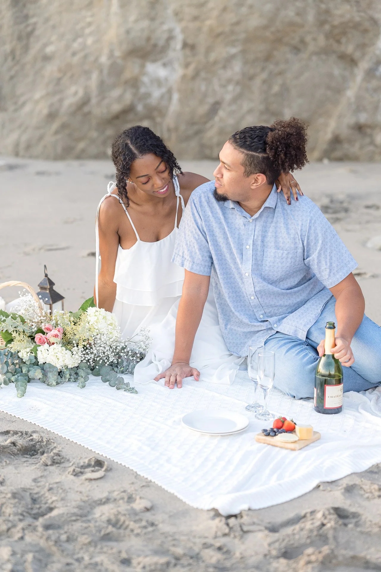 Summer Engagement Photos in Malibu featuring a couple enjoying a romantic beach picnic with champagne, fresh fruit, and flowers beside the coastal cliffs.