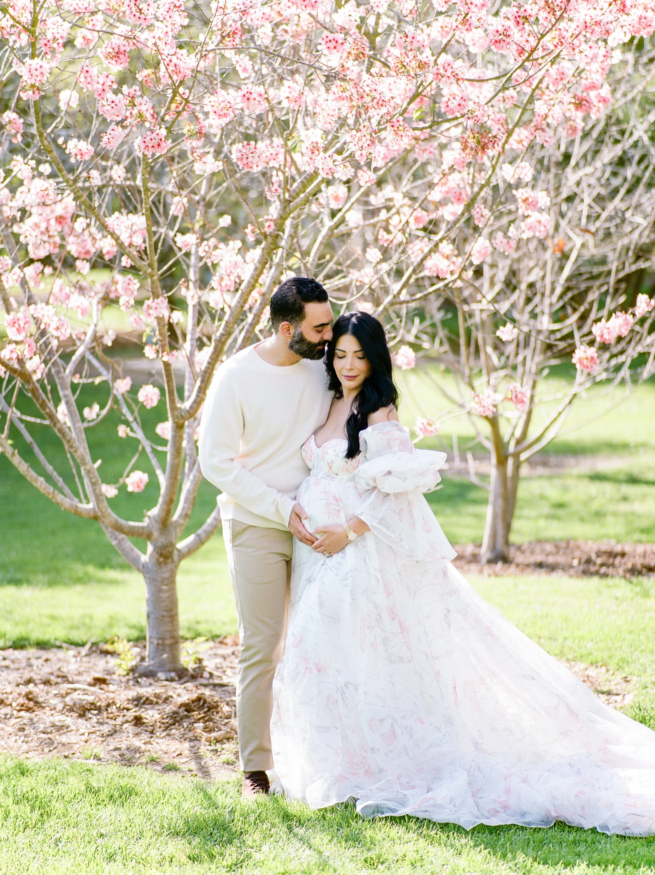 Cherry blossom Maternity photos of an expecting couple standing beneath blooming pink trees, with the mother in a flowing floral gown and her partner gently holding her baby bump in soft spring light.