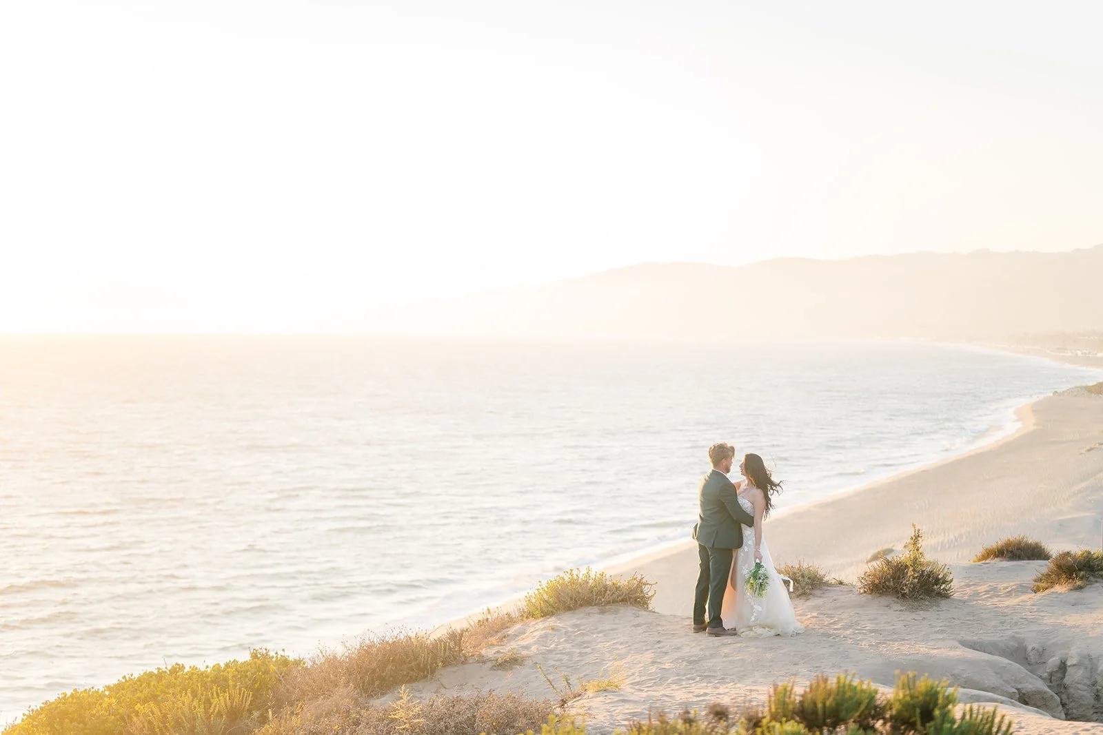 A bride and groom stand together on a sunlit cliff at Point Dume in Malibu, overlooking the ocean at sunset. Soft golden light washes over the coastline as the couple embraces, with gentle waves and distant mountains fading into the warm haze.