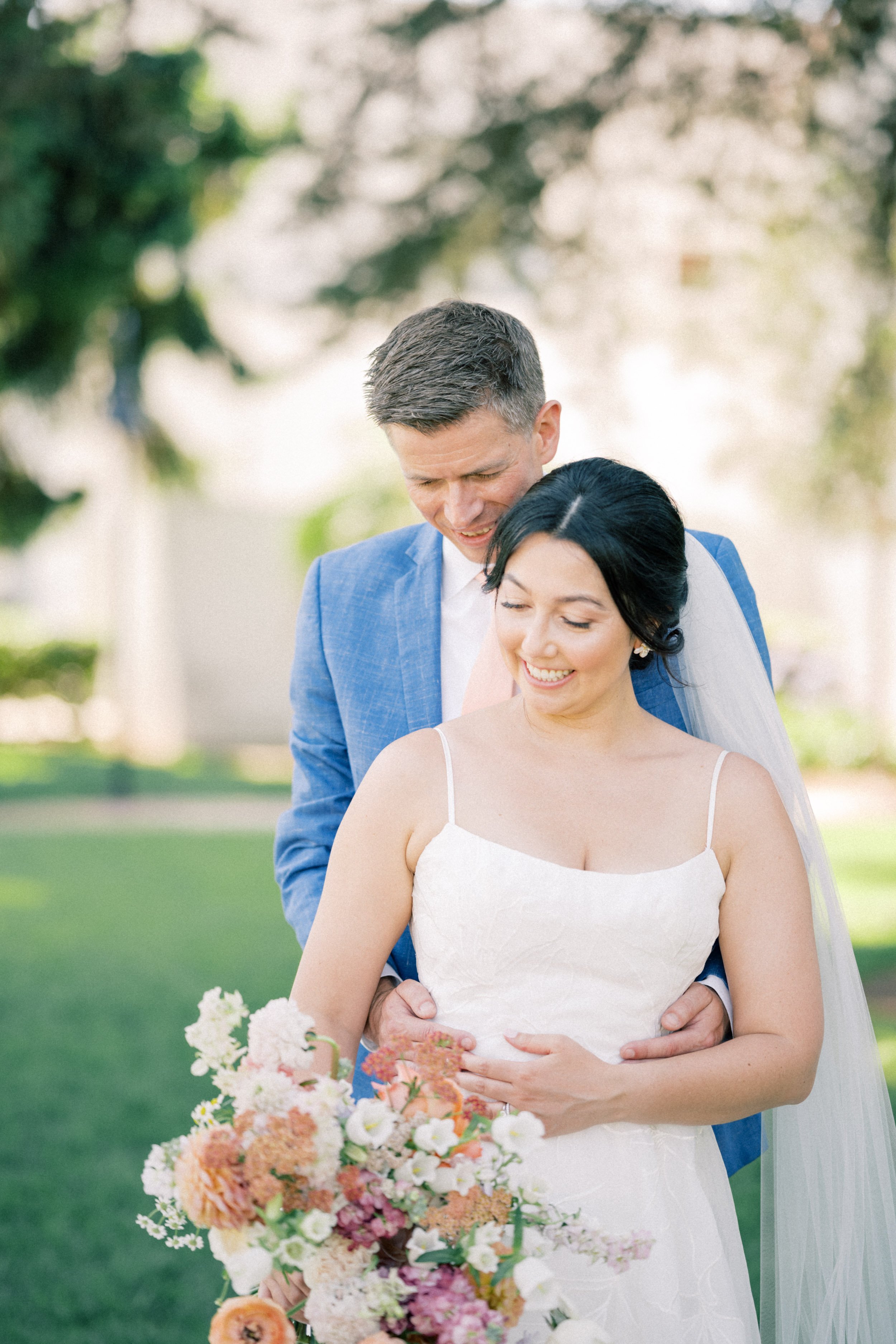 Malibu Photographer captures a romantic wedding portrait of a bride in a white gown holding a pastel floral bouquet as her groom embraces her from behind in a lush garden setting.