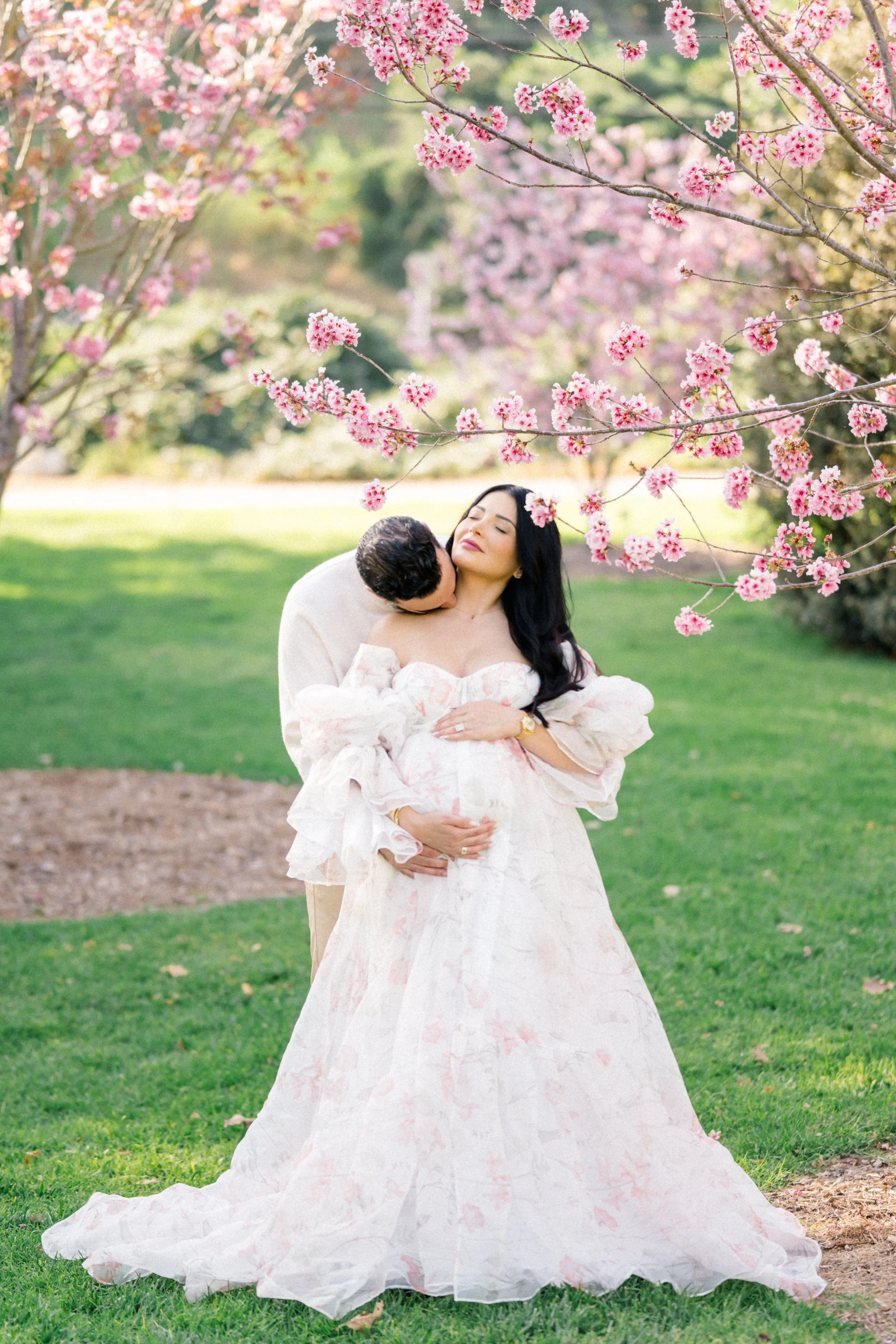 Cherry blossom Maternity photos of an expecting couple beneath blooming pink trees, with the partner embracing from behind and kissing the mother’s shoulder as they cradle her baby bump in soft spring light.