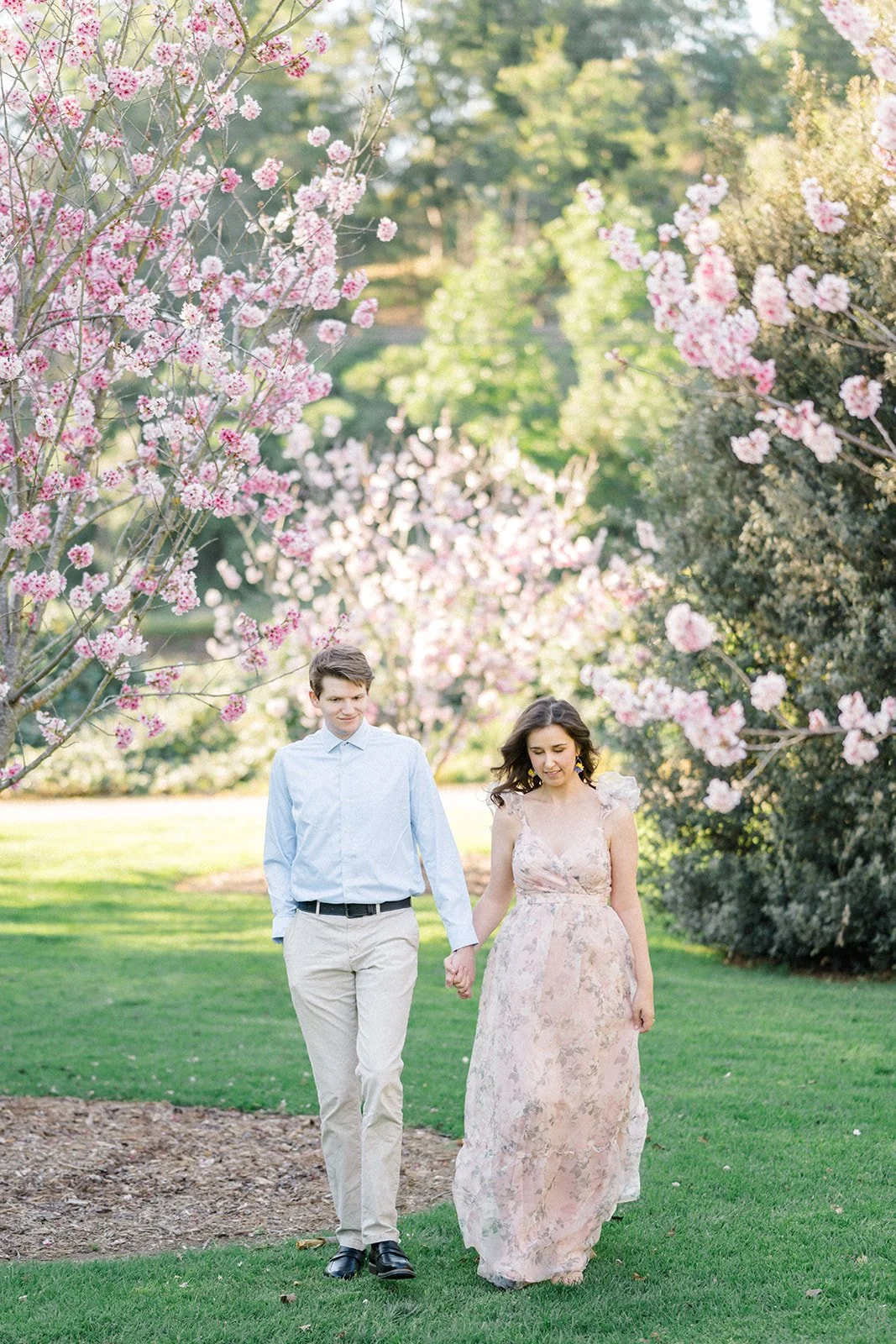 Cherry Blossom Photoshoot ideas with a couple standing close together beneath blooming cherry blossom trees, soft pink flowers framing the scene while sunlight filters through the branches for a romantic, airy feel