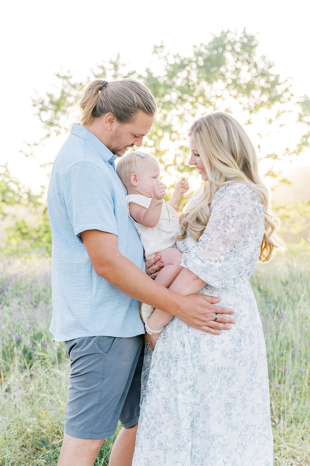 Young family shares a tender moment in a sunlit field, with the father holding their baby girl as both parents smile warmly at her, surrounded by soft golden light and wildflowers.