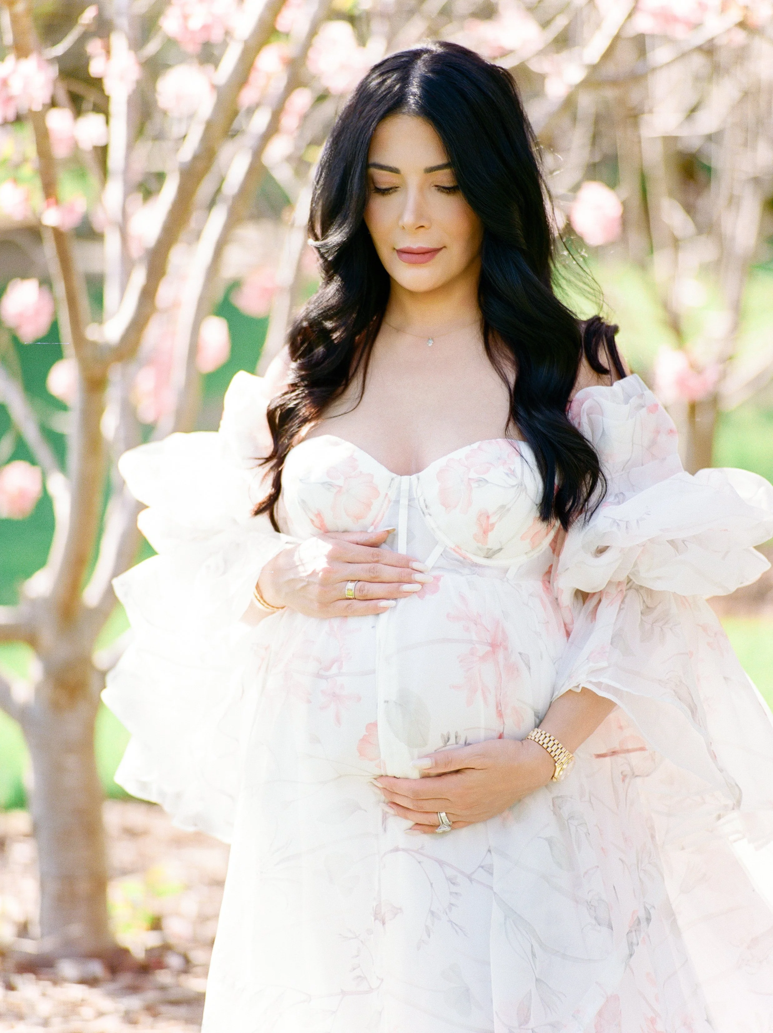Cherry blossom Maternity photos of an expecting mother in a soft floral gown gently holding her baby bump, surrounded by blooming pink trees and warm natural light.