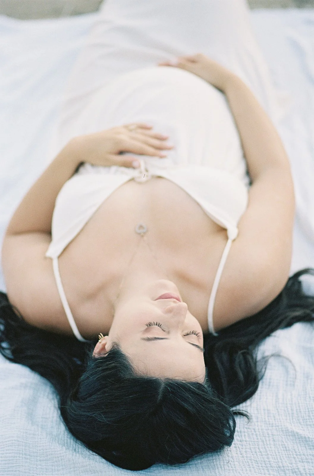 Summer Maternity Photos of an expecting mother lying on a white blanket outdoors in a soft cream dress with natural light and a relaxed editorial feel.