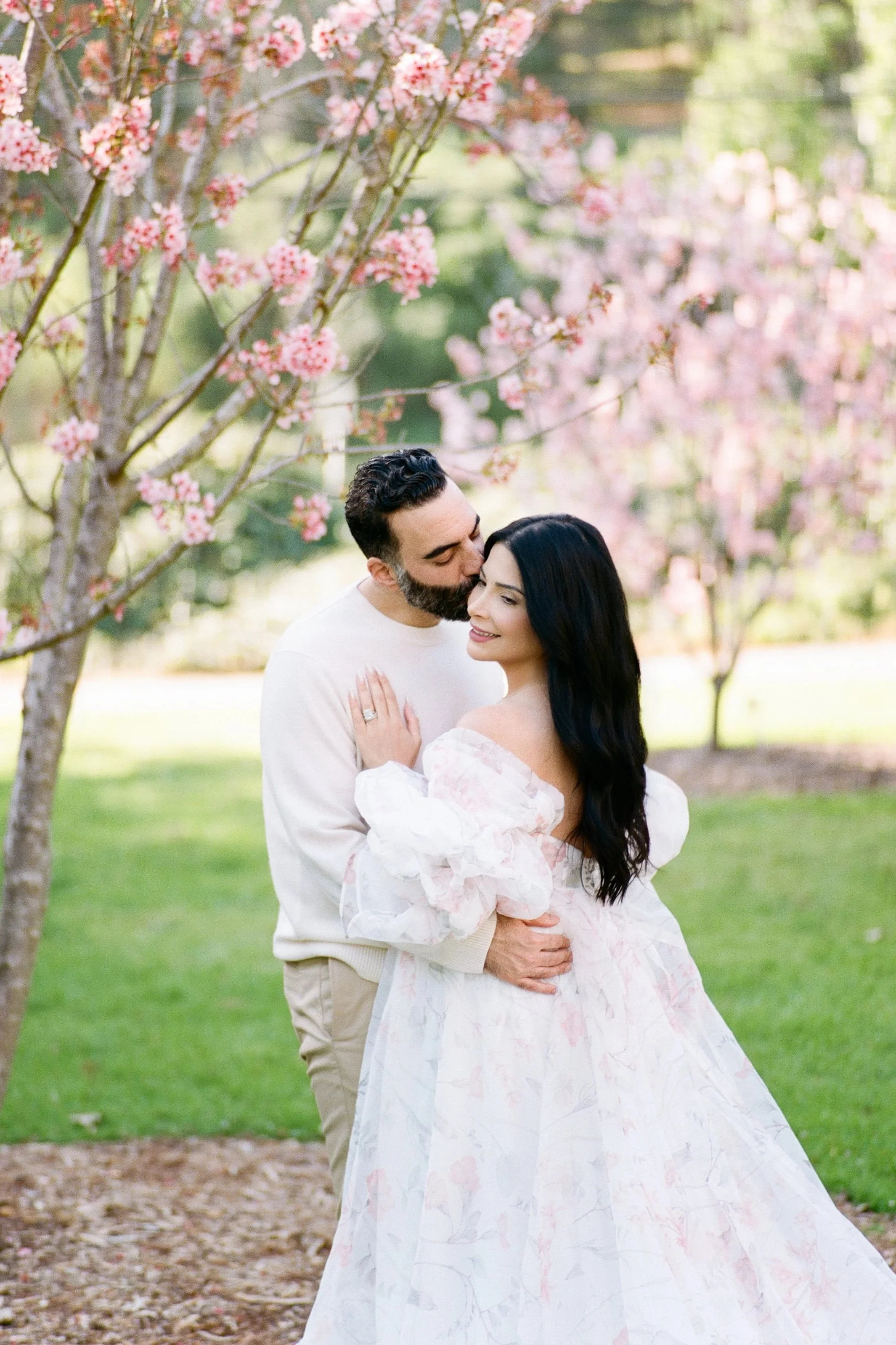 Cherry blossom Maternity photos of an expecting couple embracing beneath blooming pink trees, with the partner gently kissing the mother’s temple as they hold her baby bump in soft, natural spring light.