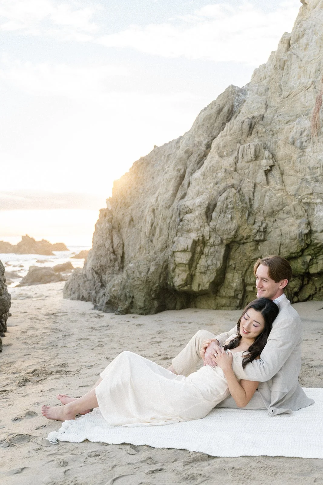 Summer Engagement Photos in Malibu featuring a couple cuddling on a blanket beneath dramatic beach cliffs with the ocean and golden sunset in the background.