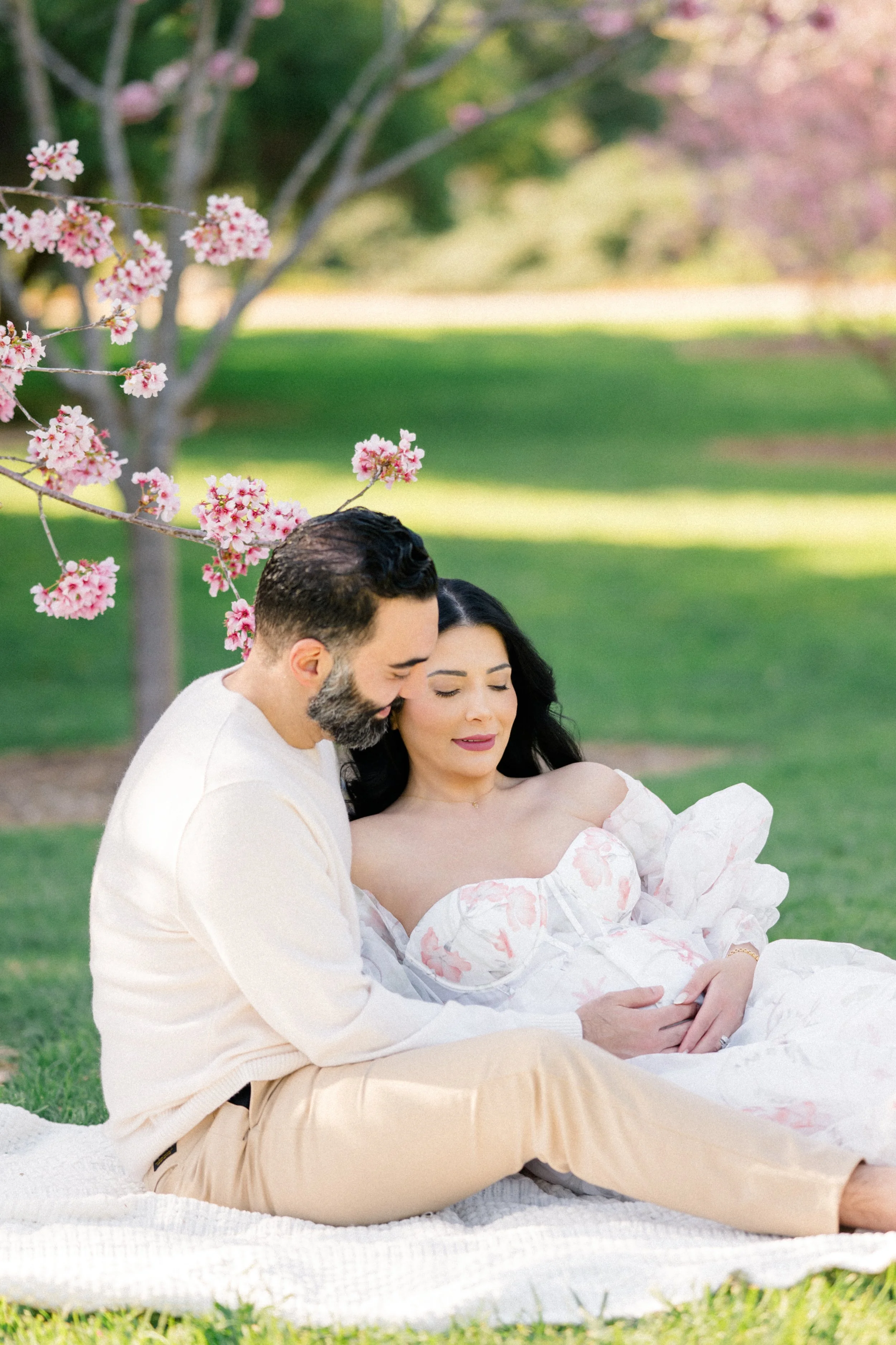 Cherry blossom Maternity photos of an expecting couple sitting on a blanket beneath blooming pink trees, with the partner gently embracing the mother as they cradle her baby bump in soft spring light.