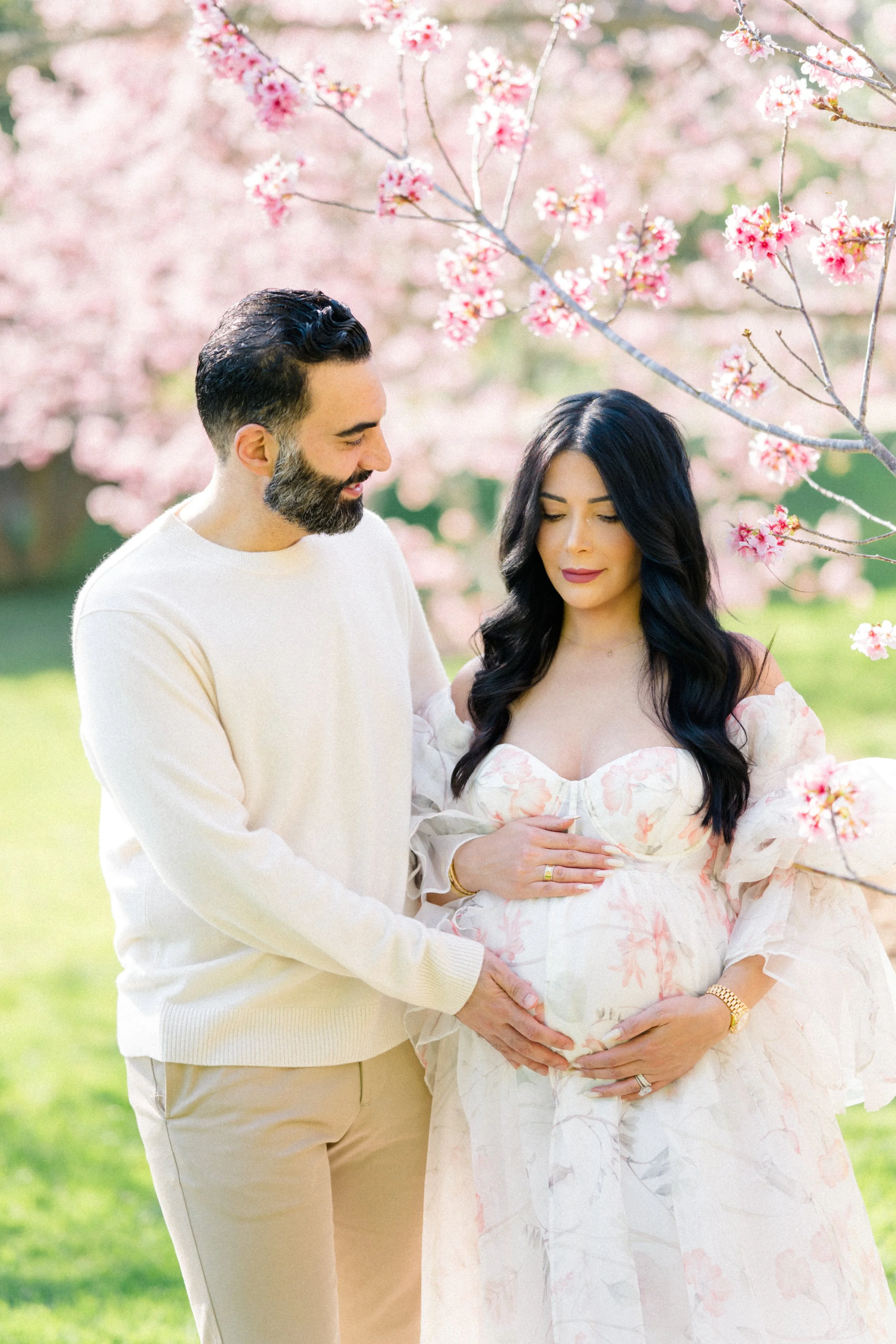 Cherry blossom Maternity photos of an expecting couple standing beneath blooming pink trees, with the partner gently holding the mother’s baby bump as they share a soft, intimate moment in natural light.