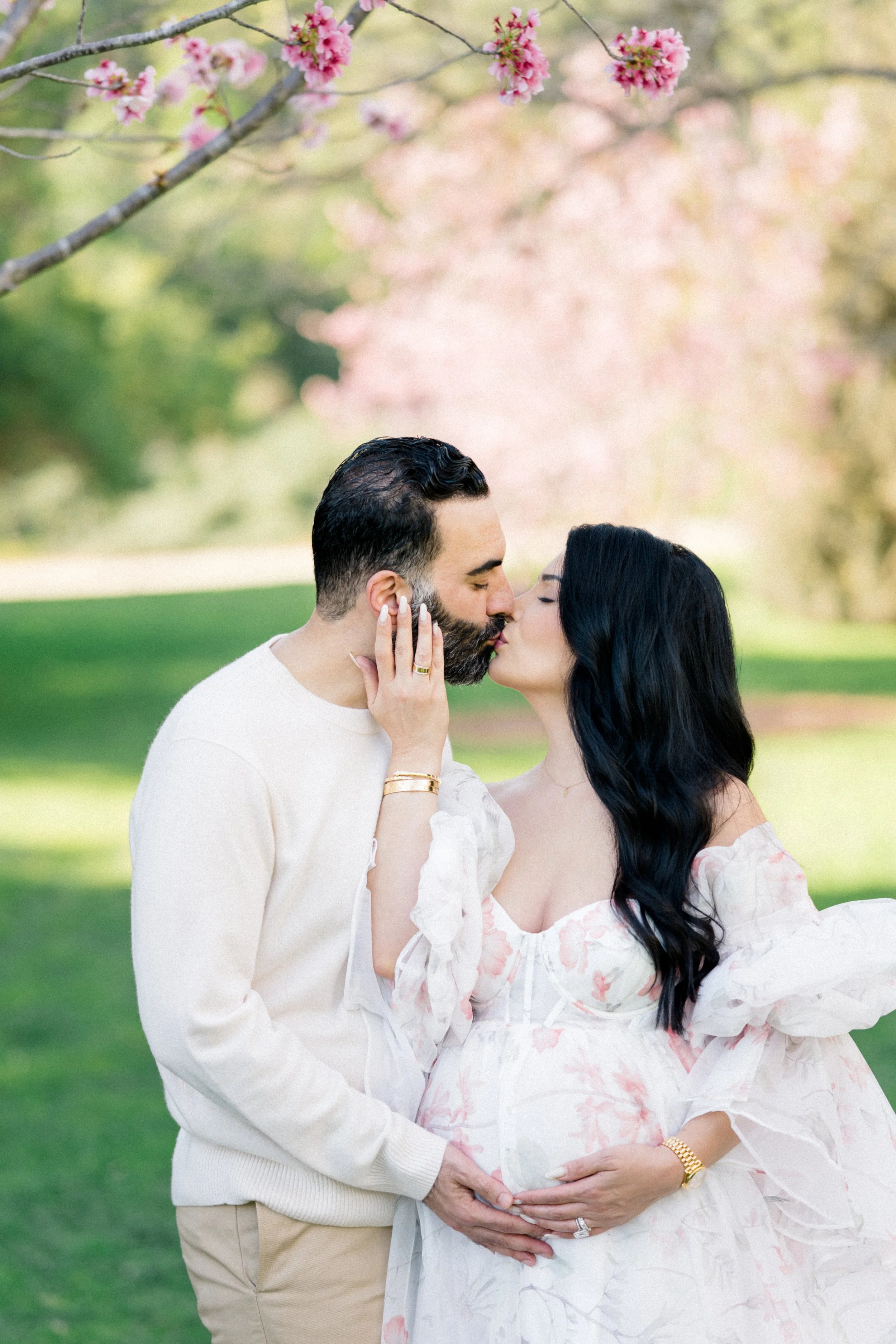 Cherry blossom Maternity photos of an expecting couple sharing a kiss beneath soft pink blossoms, gently cradling the baby bump in warm, natural spring light.
