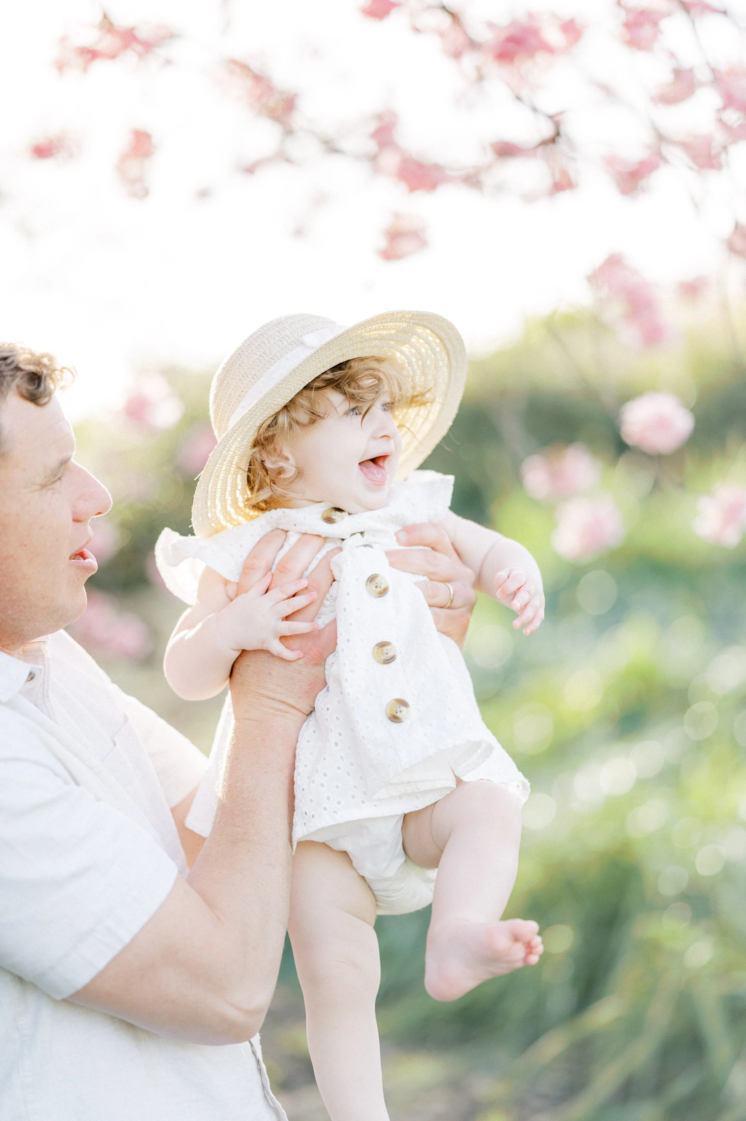 Cherry Blossom Photoshoot ideas featuring a candid family moment of a parent lifting a smiling baby wearing a sun hat, with soft pink cherry blossoms glowing in the background in warm spring light