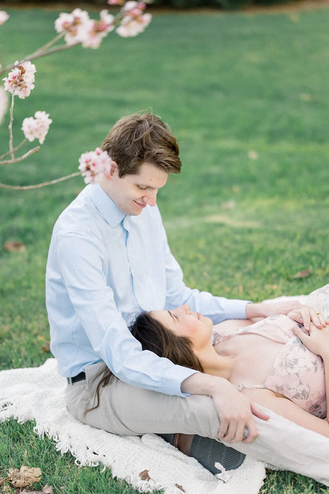 Cherry Blossom Photoshoot ideas featuring a couple relaxing on a blanket beneath blooming cherry blossom branches, with the woman laying across her partner’s lap as they share a quiet, intimate moment in soft spring light