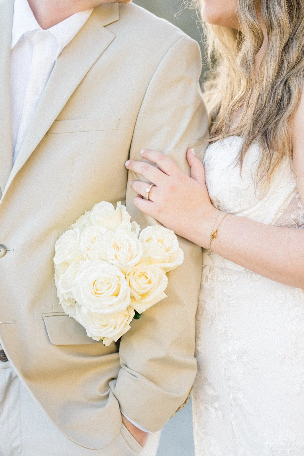 Valentines Day Photoshoot Ideas shown through a close-up of a couple in neutral wedding attire with a bouquet of white roses and an engagement ring during a beach session.