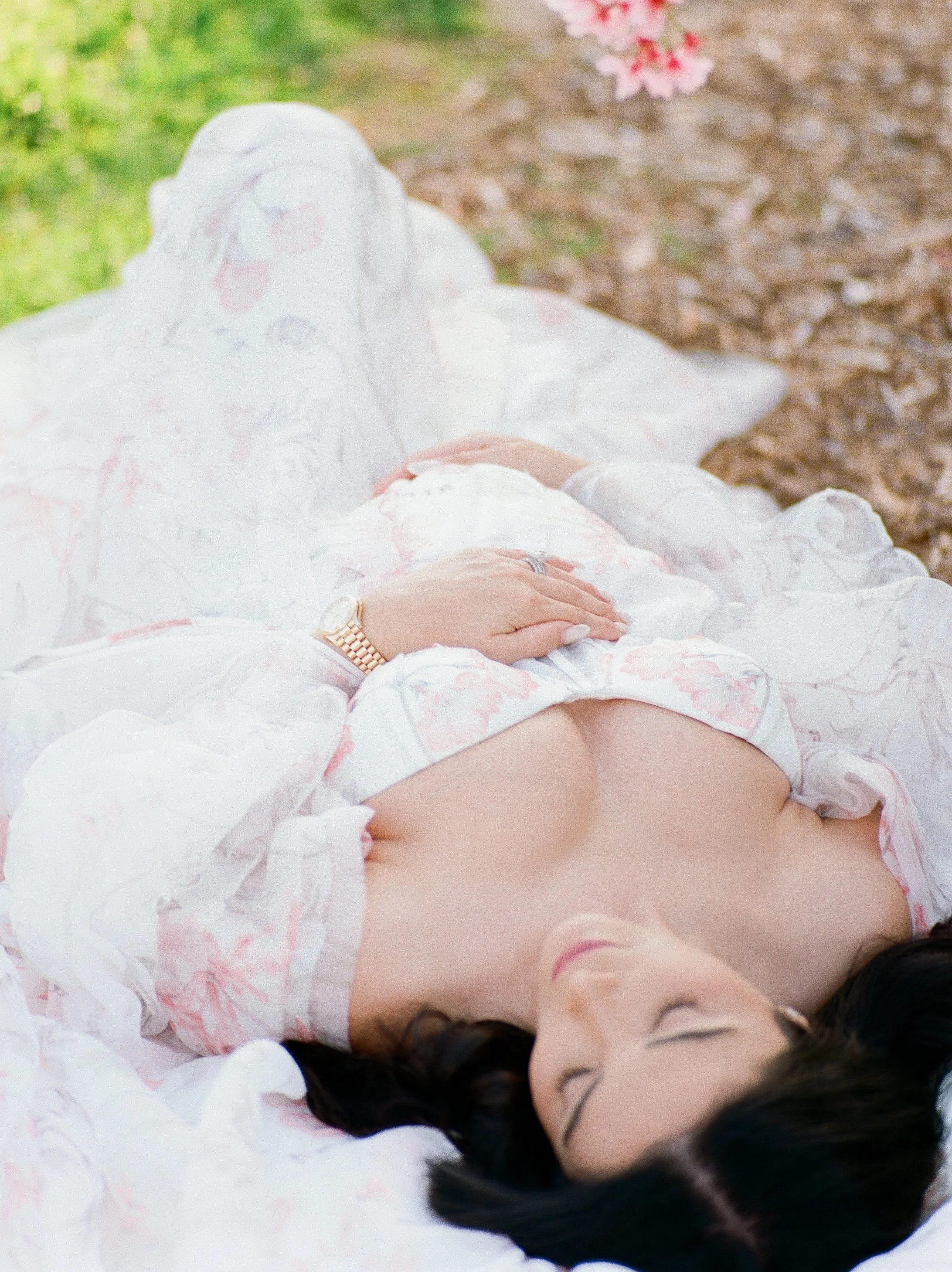 Cherry blossom Maternity photos close-up of an expecting mother lying down in a soft floral gown, gently resting her hands on her baby bump in warm, natural spring light.