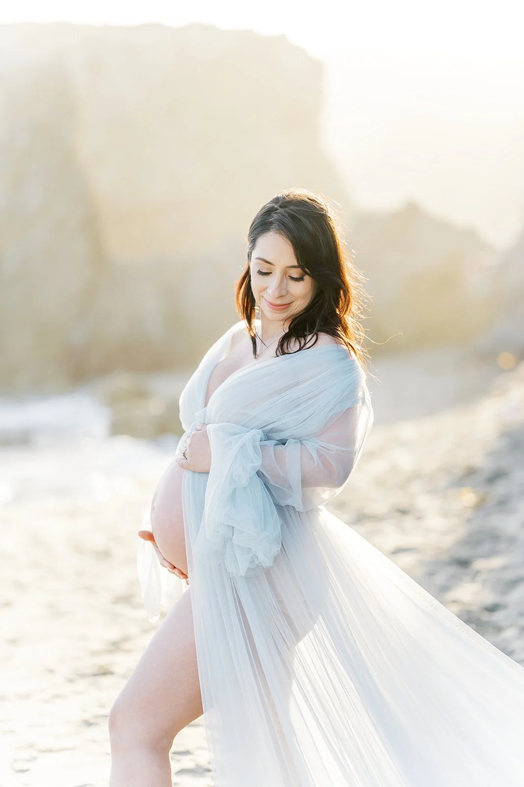 Summer Maternity Photos of an expecting mother in a flowing sheer gown standing on the beach during golden hour with soft ocean light and coastal cliffs in the background.
