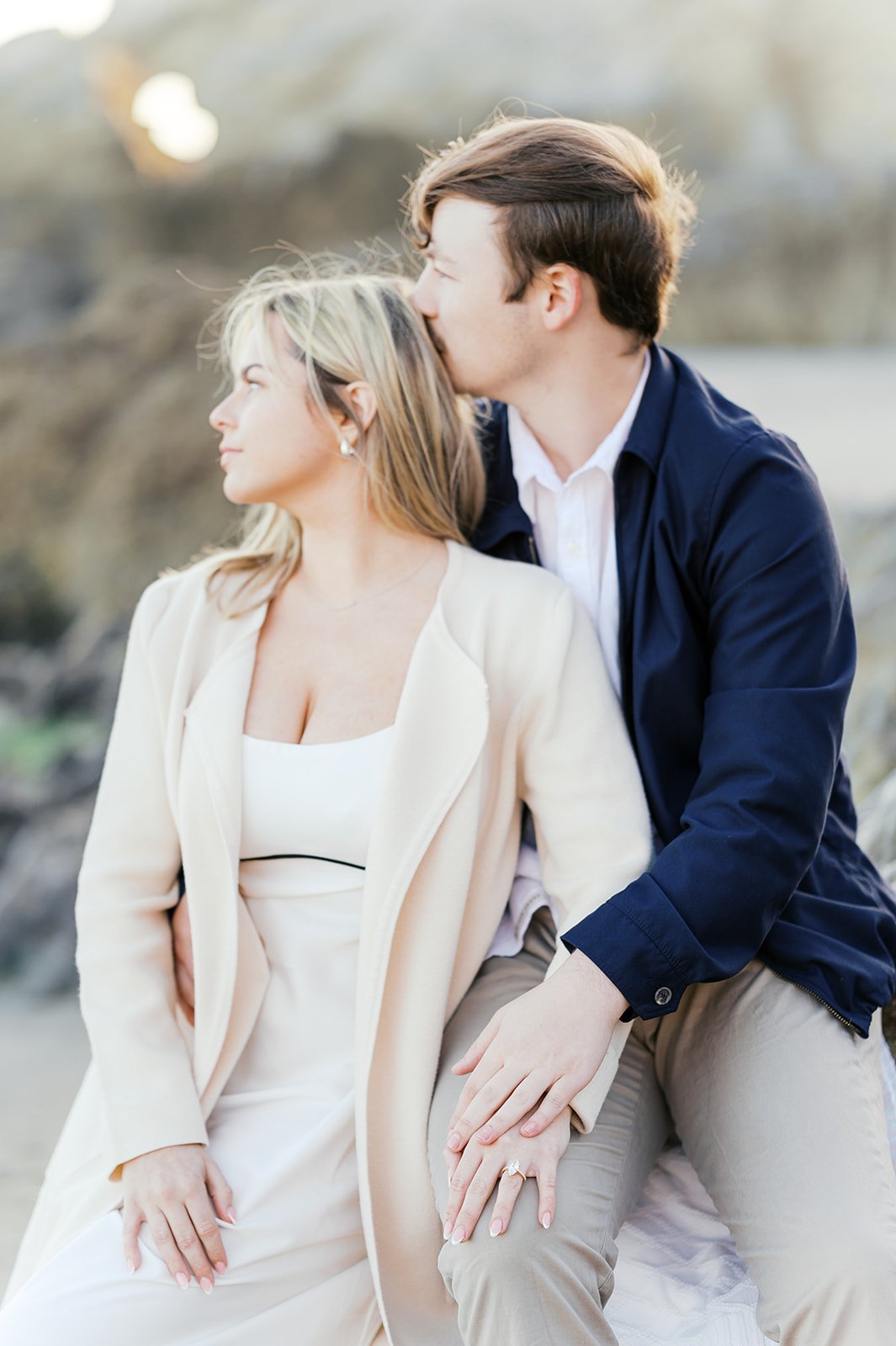 Summer Engagement Photos in Malibu featuring an engaged couple sitting together outdoors as the groom kisses his fiancée’s head and she shows her engagement ring.