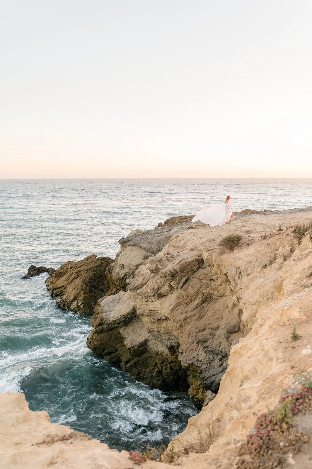 Pregnant woman in a flowing gown stands on a rocky cliff overlooking the ocean at sunset, waves crashing below and soft pastel light illuminating the coastal landscape.