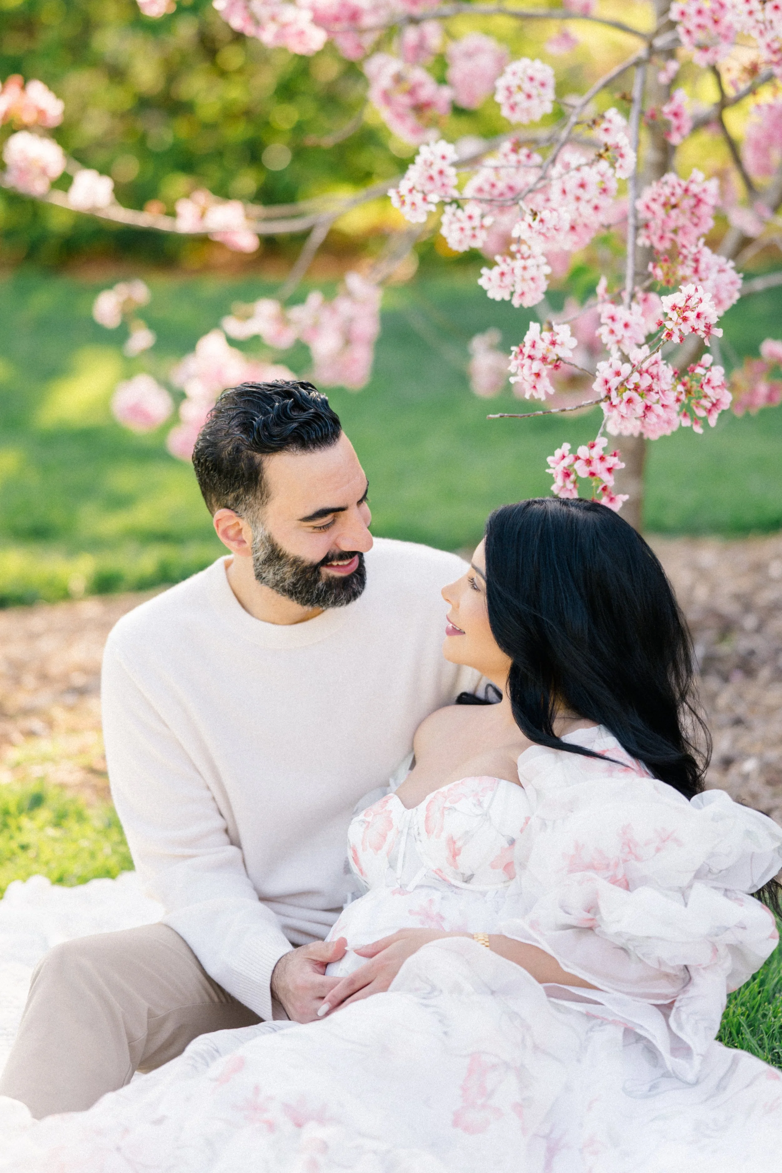 Cherry blossom Maternity photos of an expecting couple sitting together beneath blooming pink trees, smiling at each other while gently holding the baby bump in soft spring light.