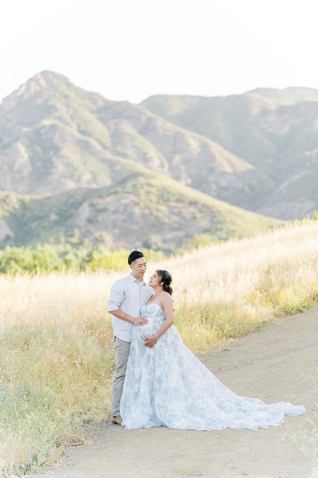 Summer Maternity Photos of an expecting couple embracing on a mountain trail surrounded by golden fields and soft California sunset light.