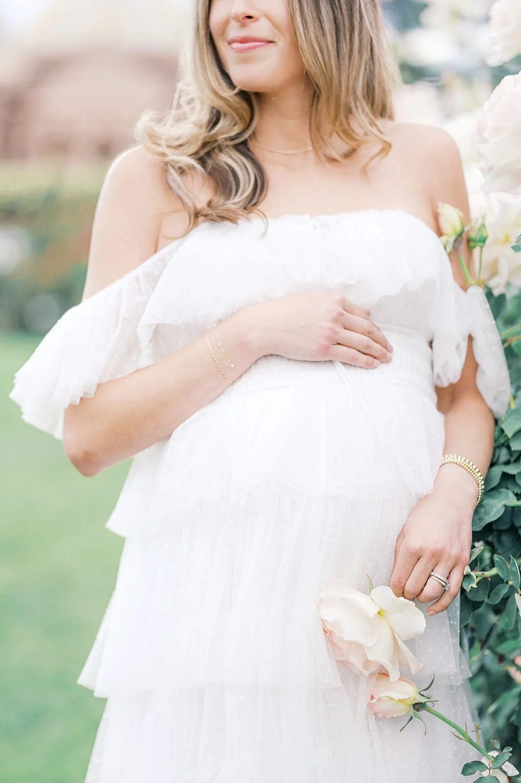 Close-up Summer Maternity Photos of an expecting mother in a soft white off-the-shoulder dress holding roses in a blooming summer garden.