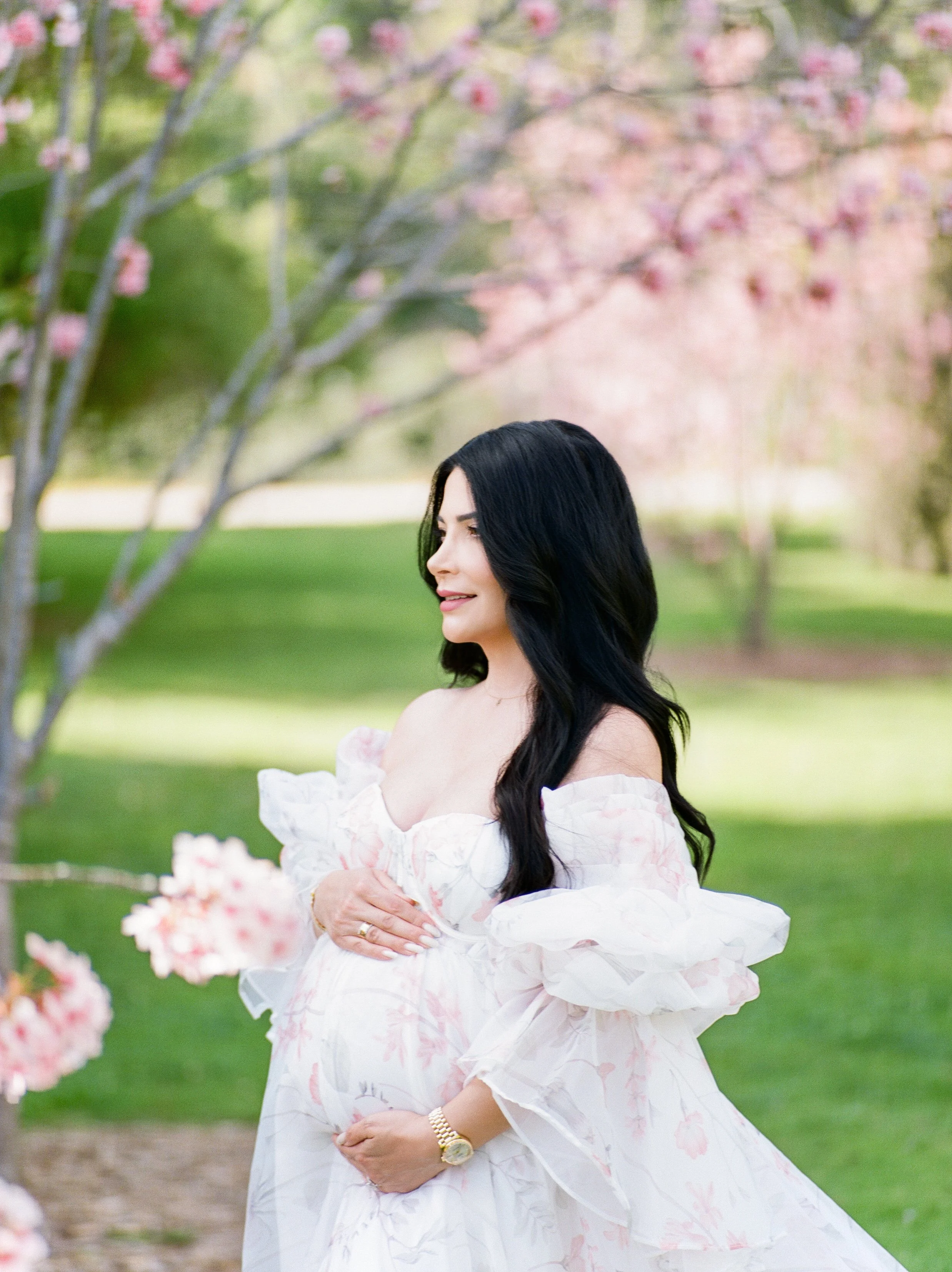 Cherry blossom Maternity photos of an expecting mother in profile, gently holding her baby bump in a flowing floral gown, surrounded by soft pink blossoms in natural spring light.