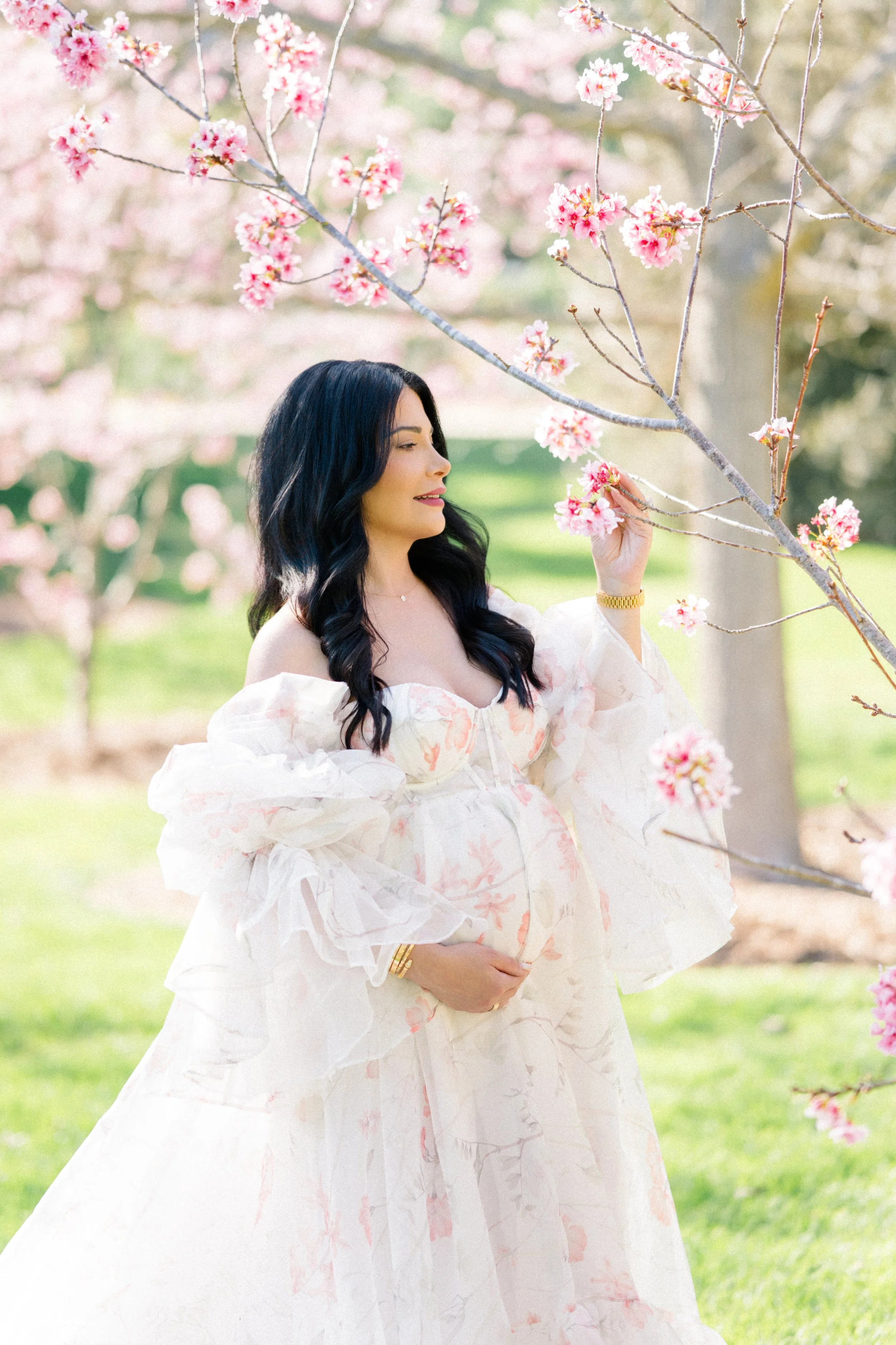 Cherry blossom Maternity photos of an expecting mother in a flowing floral gown gently holding her baby bump while reaching toward blooming pink blossoms in soft spring light.