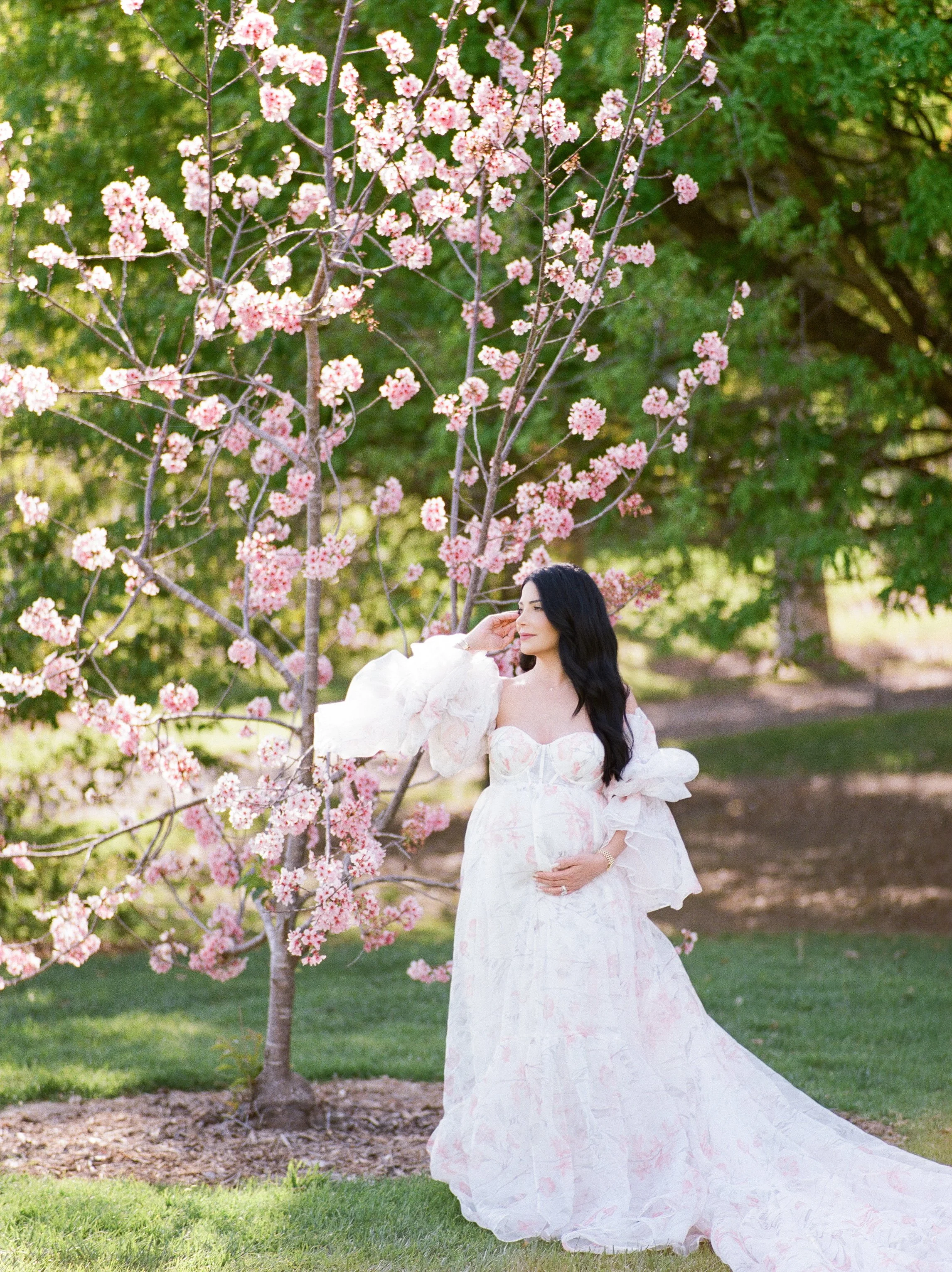 Cherry blossom Maternity photos of an expecting mother standing beside a blooming pink tree, wearing a flowing floral gown and gently holding her baby bump in soft, natural spring light.
