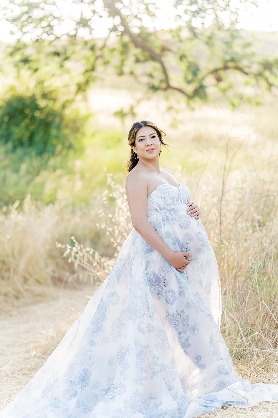 Summer Maternity Photos of an expecting mother in a soft blue floral gown standing in a sunlit field with golden grass and oak trees in the background.
