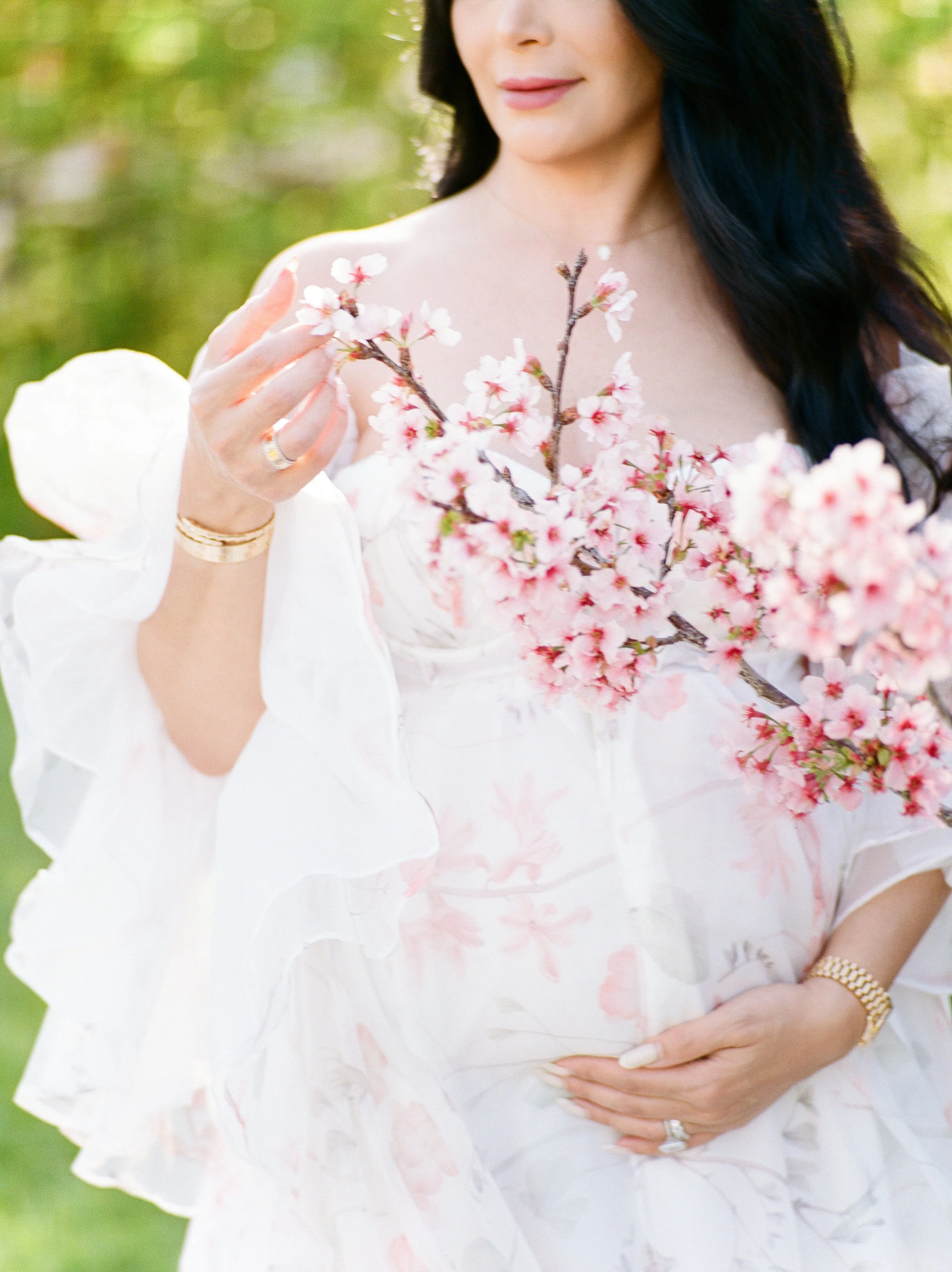 Cherry blossom Maternity photos close-up of an expecting mother in a soft floral gown gently holding her baby bump while touching delicate pink blossoms in warm, natural spring light.